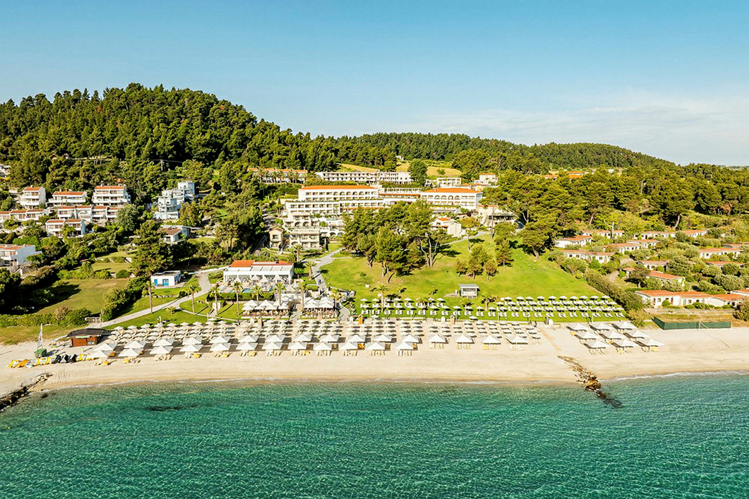 Luftfoto af en ferieresort ved stranden med liggestole og parasoller samt omkringliggende grønne skovklædte bakker. (AI-genereret tekst)