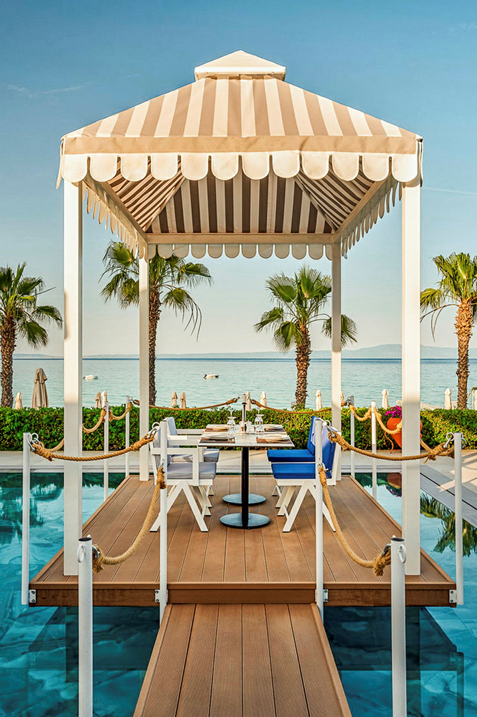 En pavillon med et spisebord og stole på en bro ved en pool. I baggrunden ses palmer og havet. (AI-genereret tekst)