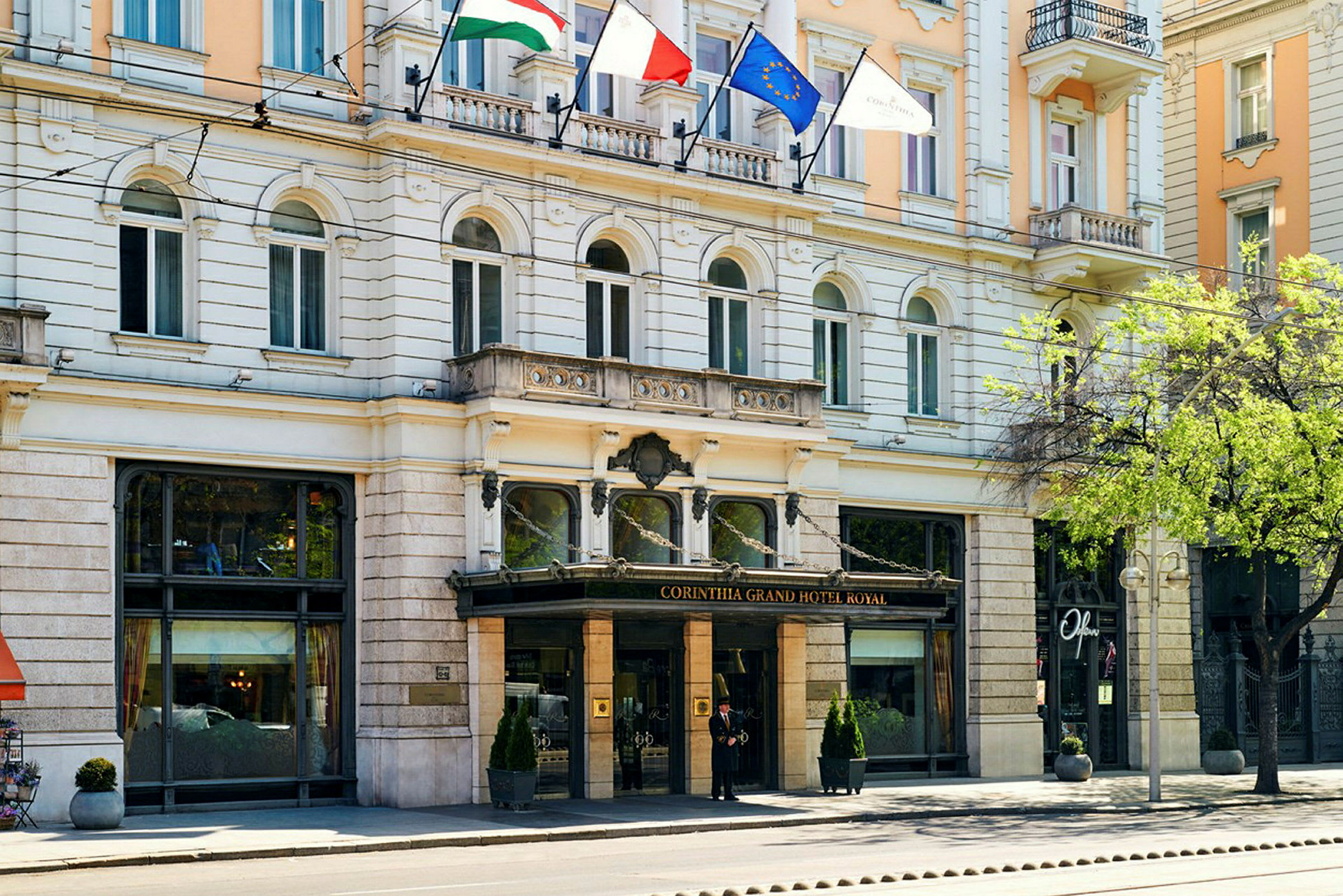 Forsiden af Corinthia Grand Hotel Royal med glasindgang og flag ovenover. (AI-genereret tekst)