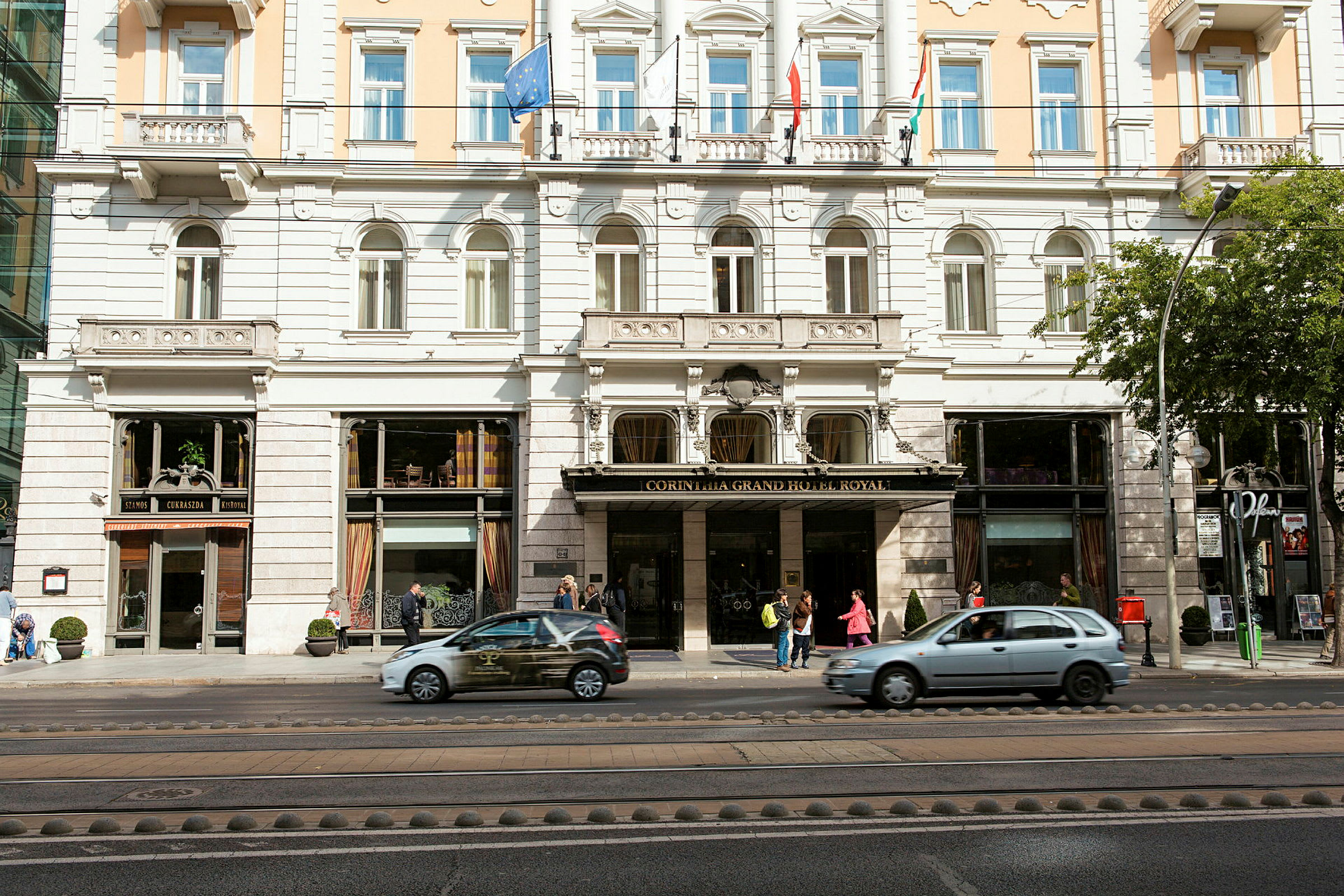 Facade af et hotel med flere etager, indvendigt synlige vinduer og indgangsområdet med mennesker og forbipasserende biler. (AI-genereret tekst)