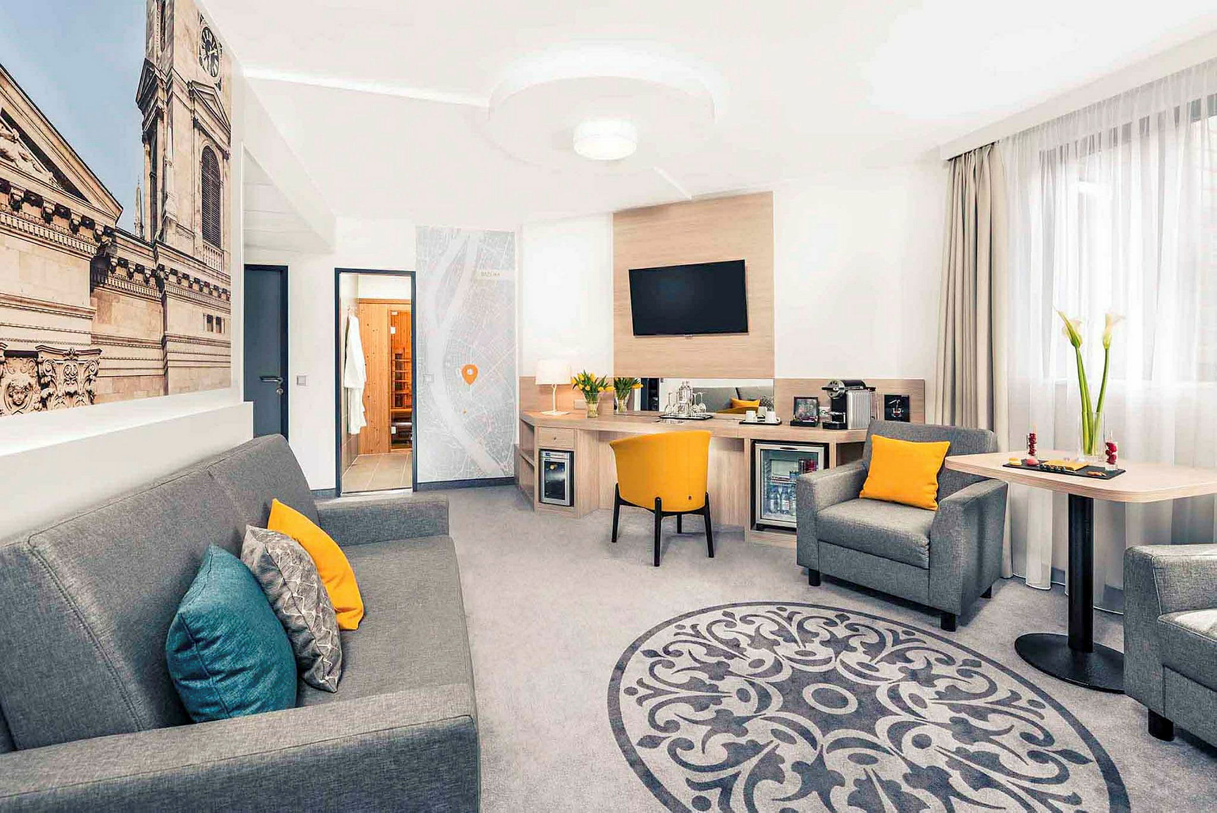 Et hotelværelse med siddegruppe, skrivebord og et stort vægbillede af en bygningsfacade. (AI-genereret tekst)
