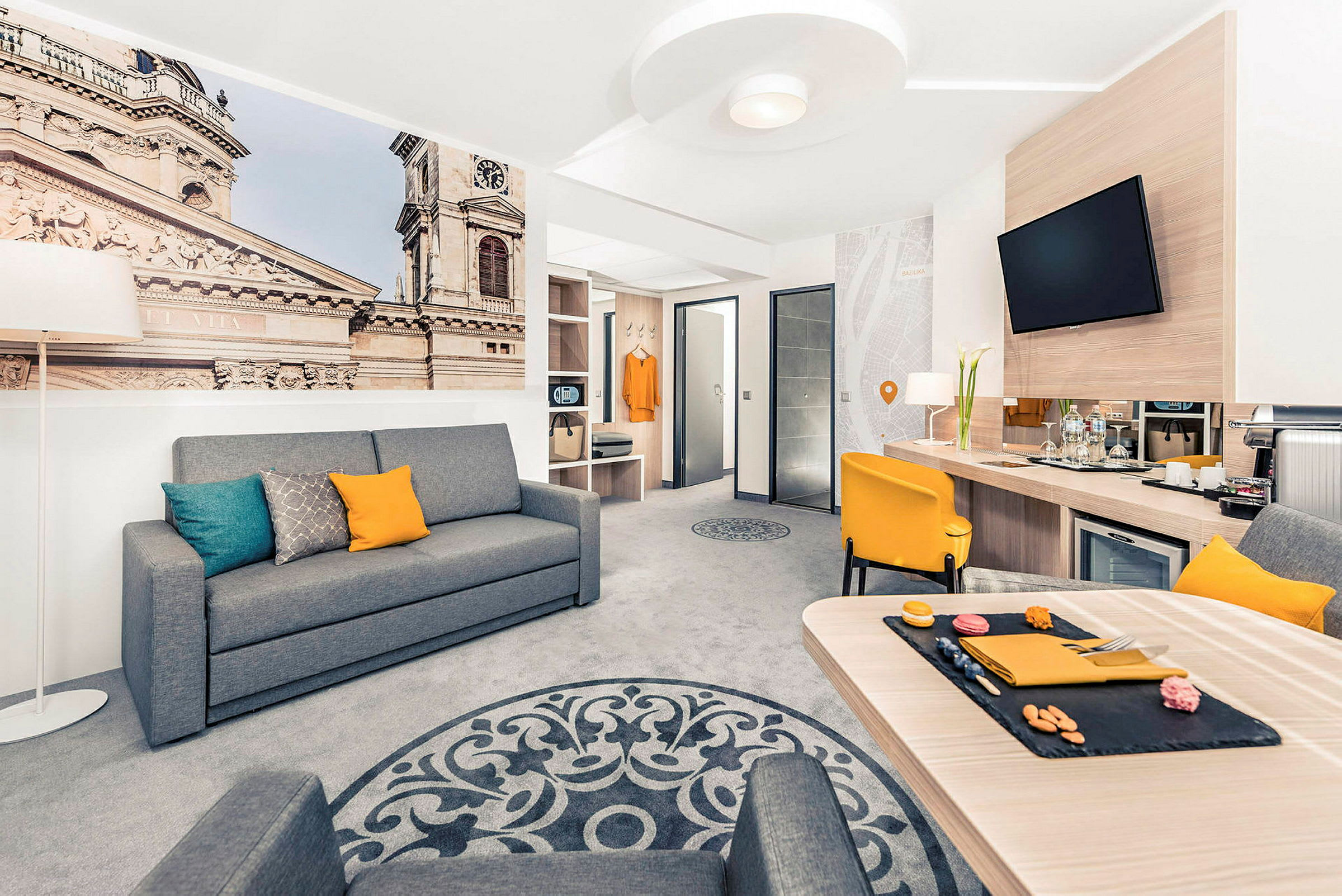 Interiør af et moderne hotelværelse med en grå sofa, gul stol og et bord, samt et stort vægmaleri der viser en bygning. (AI-genereret tekst)