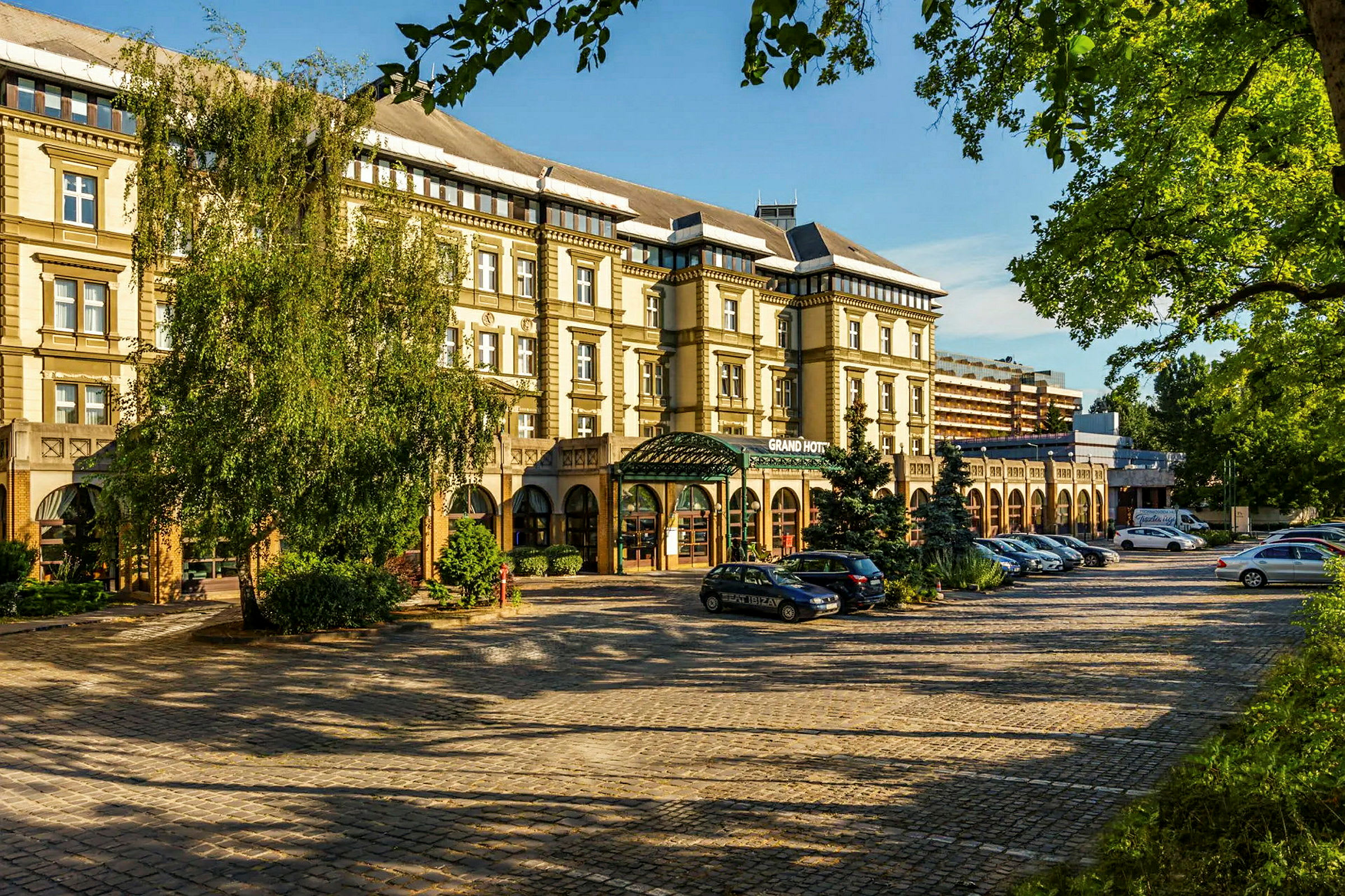 Et billede af en bygning med teksten 'Grand Hotel' på facaden, omgivet af træer og parkerede biler. (AI-genereret tekst)