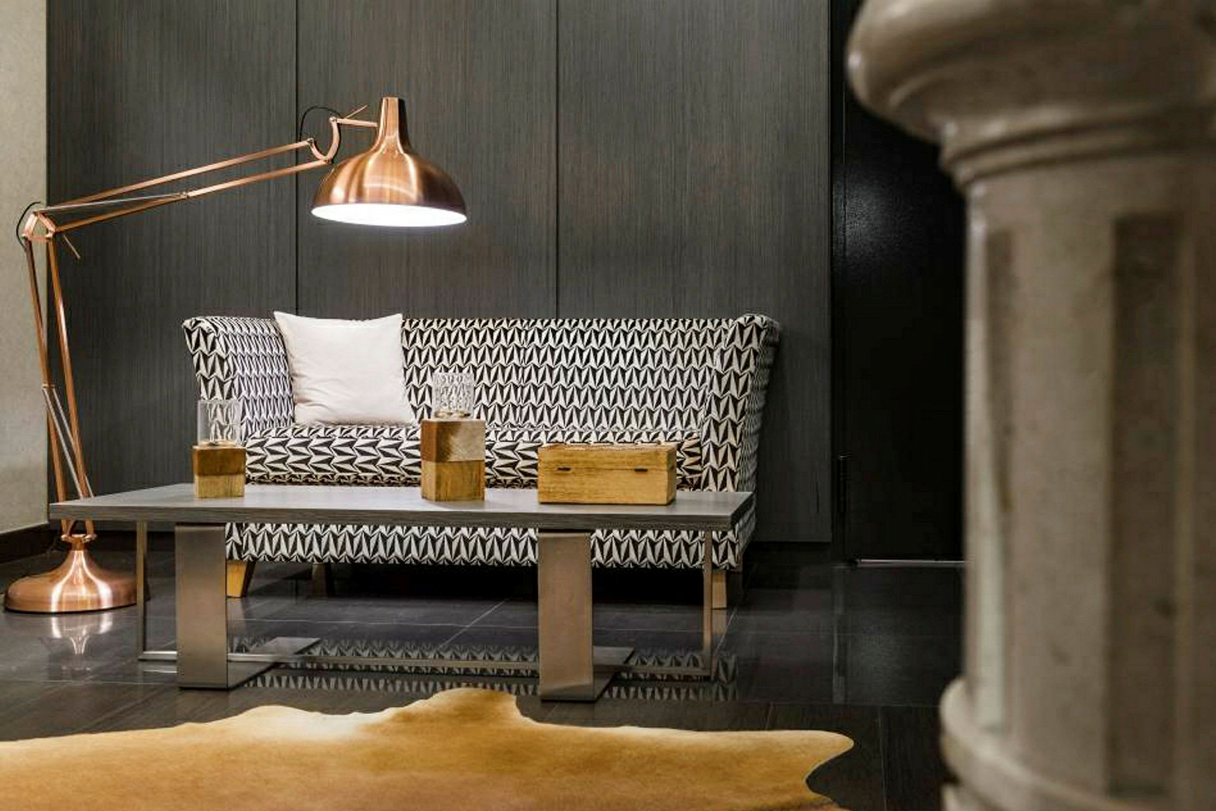 Et moderne rum med en geometrisk mønstret sofa, metalbord med træpynt og stor kobberfarvet gulvlampe. (AI-genereret tekst)