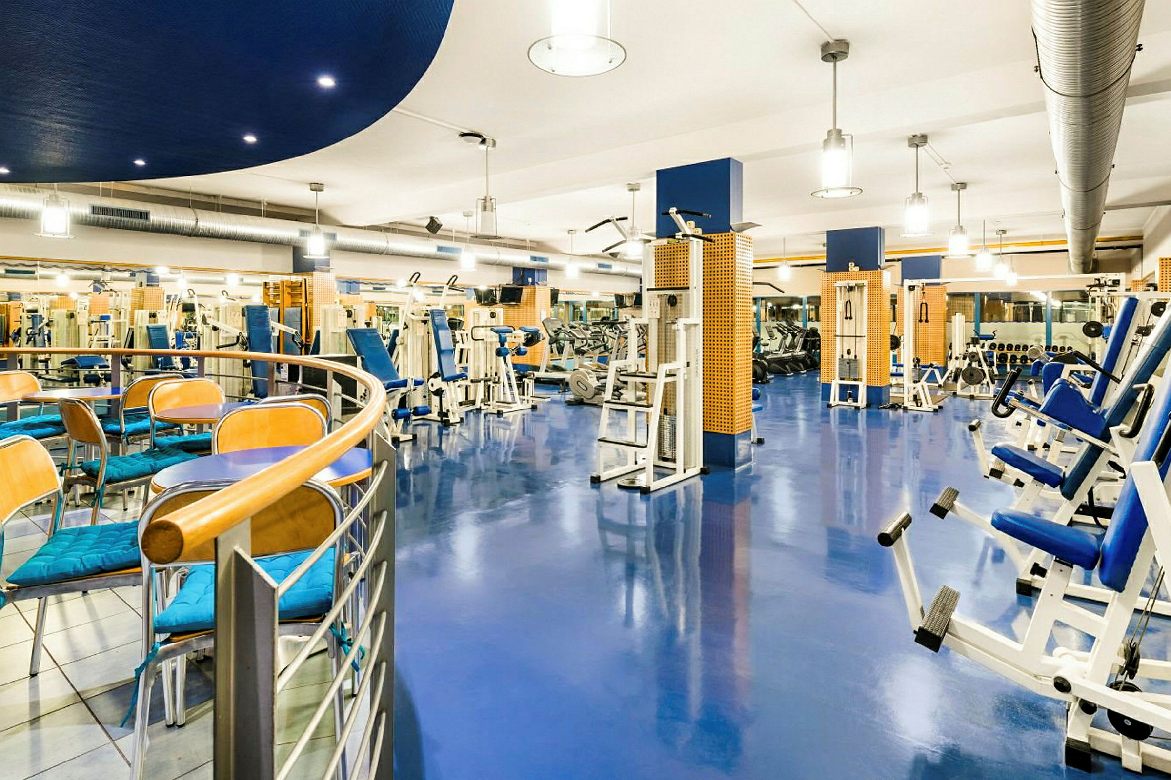 Et moderne fitnesscenter med træningsudstyr og borde med stole i forgrunden. (AI-genereret tekst)