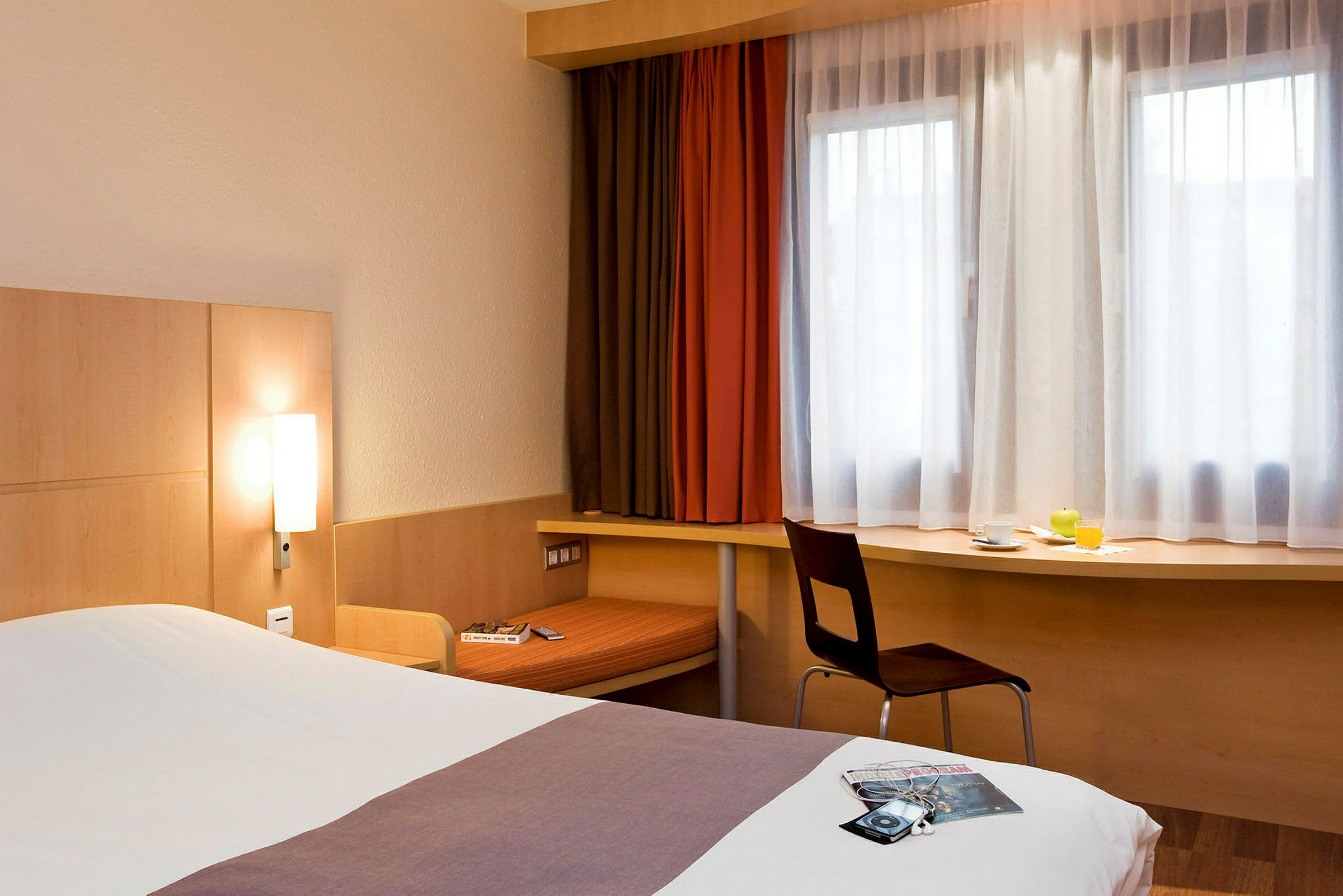Et hotelværelse med en seng, et skrivebord under vinduet og en stol. Sengen er redt, og på skrivebordet står der en kop, et glas appelsinjuice og en grøn frugt. (AI-genereret tekst)