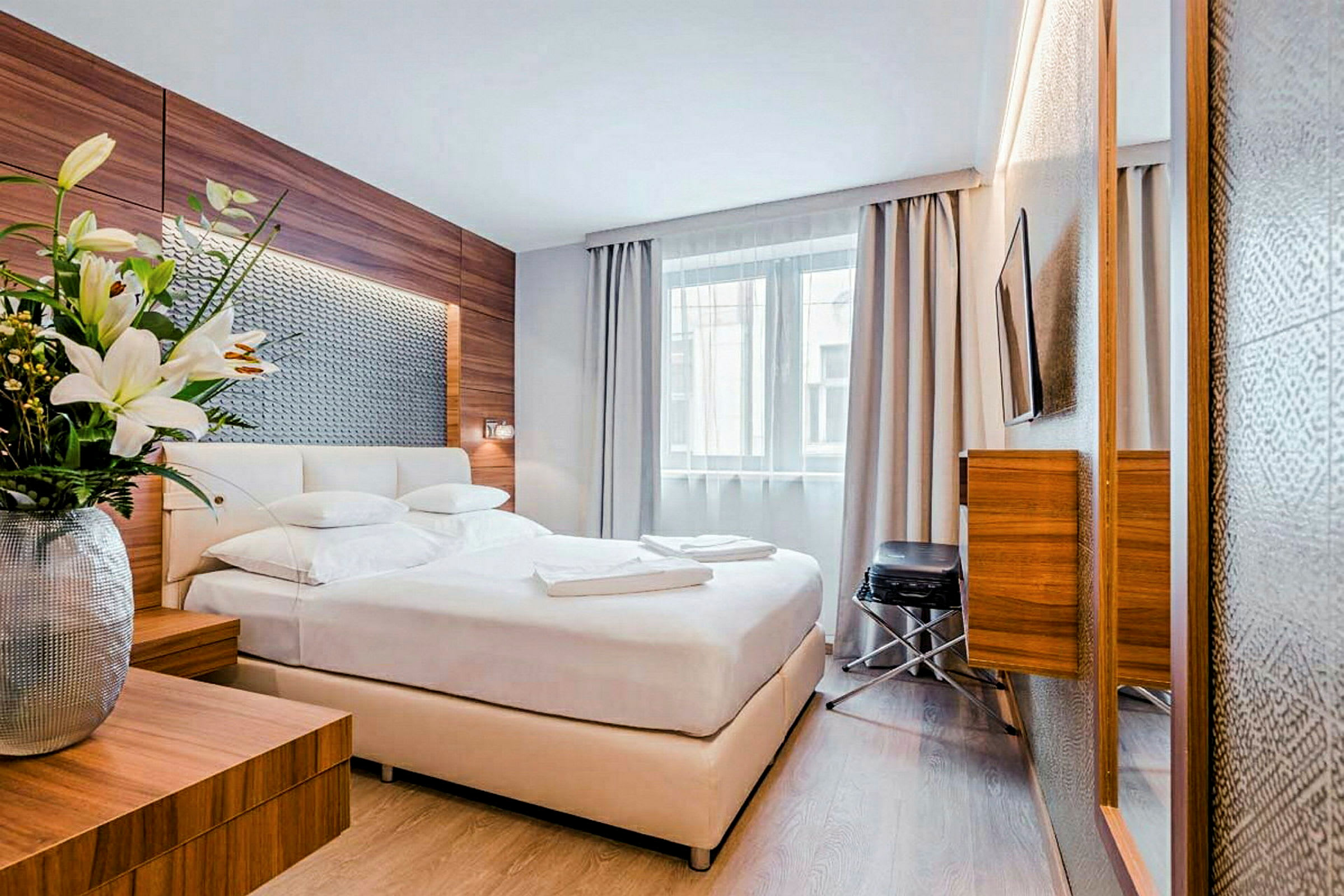 Et moderne hotelværelse med en dobbeltseng, et stort vindue med gardiner og frisk indretning med trædetaljer. En vase med blomster står på et natbord. (AI-genereret tekst)