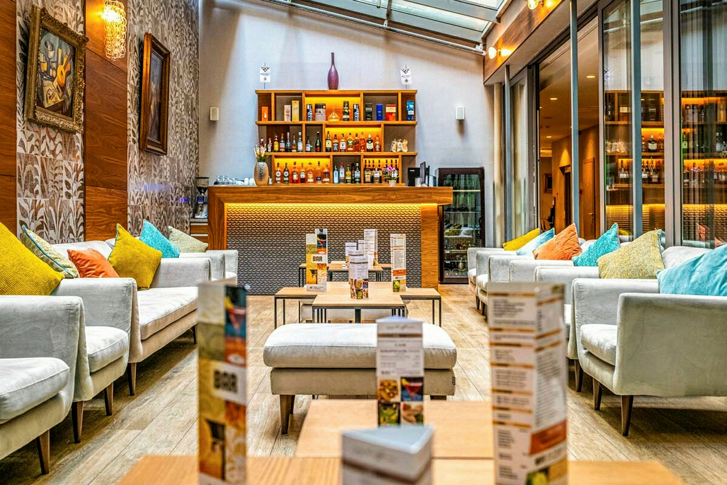 Et moderne loungeområde med sofaer og puder i forskellige farver, borde og en bar med oplyste hylder fyldt med flasker. (AI-genereret tekst)