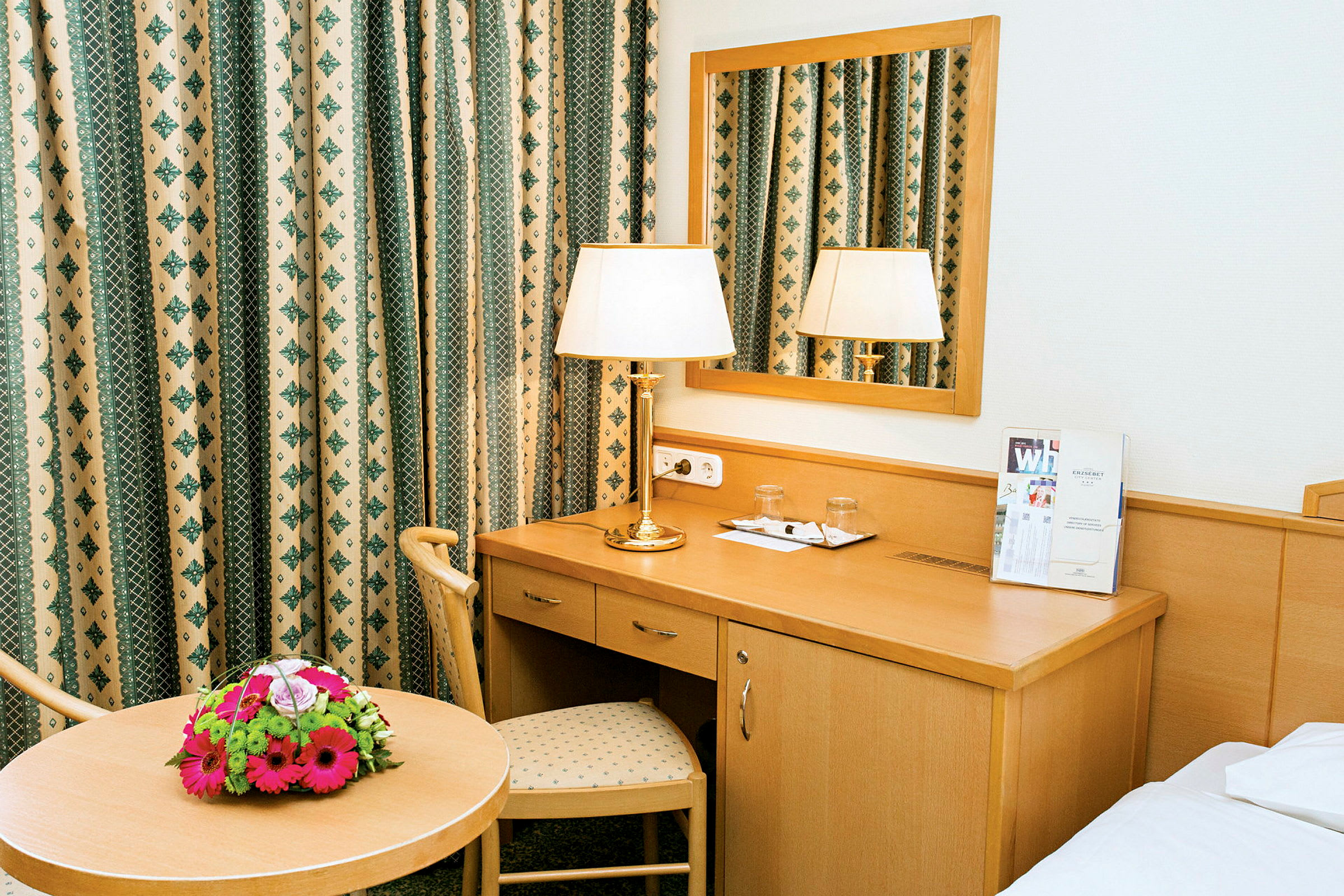 Et hotelværelse med et skrivebord, spejl, bord med blomsterbuket og lampe. (AI-genereret tekst)
