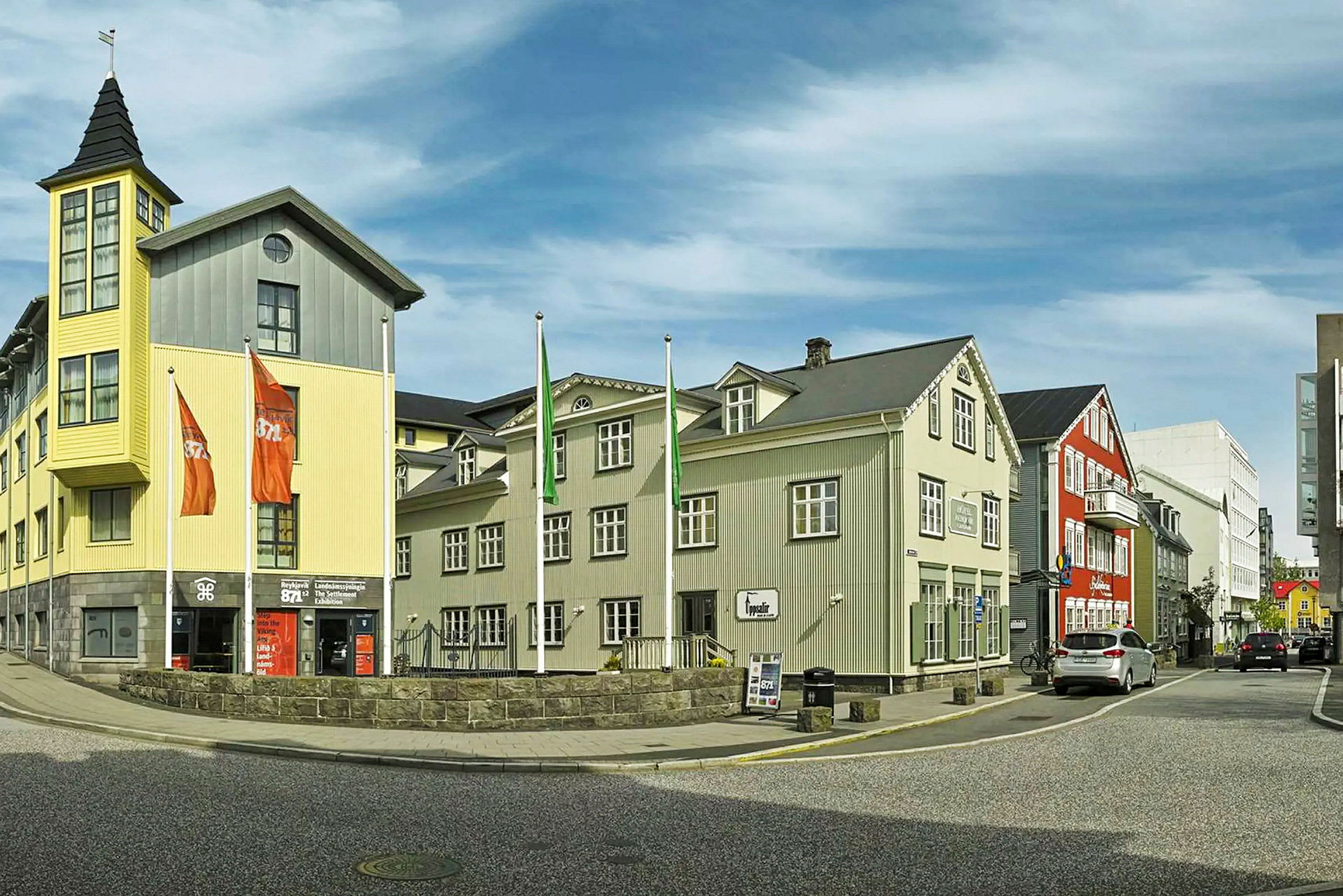 En gade med bygninger i forskellige farver, inklusive en gul bygning med et tårn og flagstænger foran. (AI-genereret tekst)