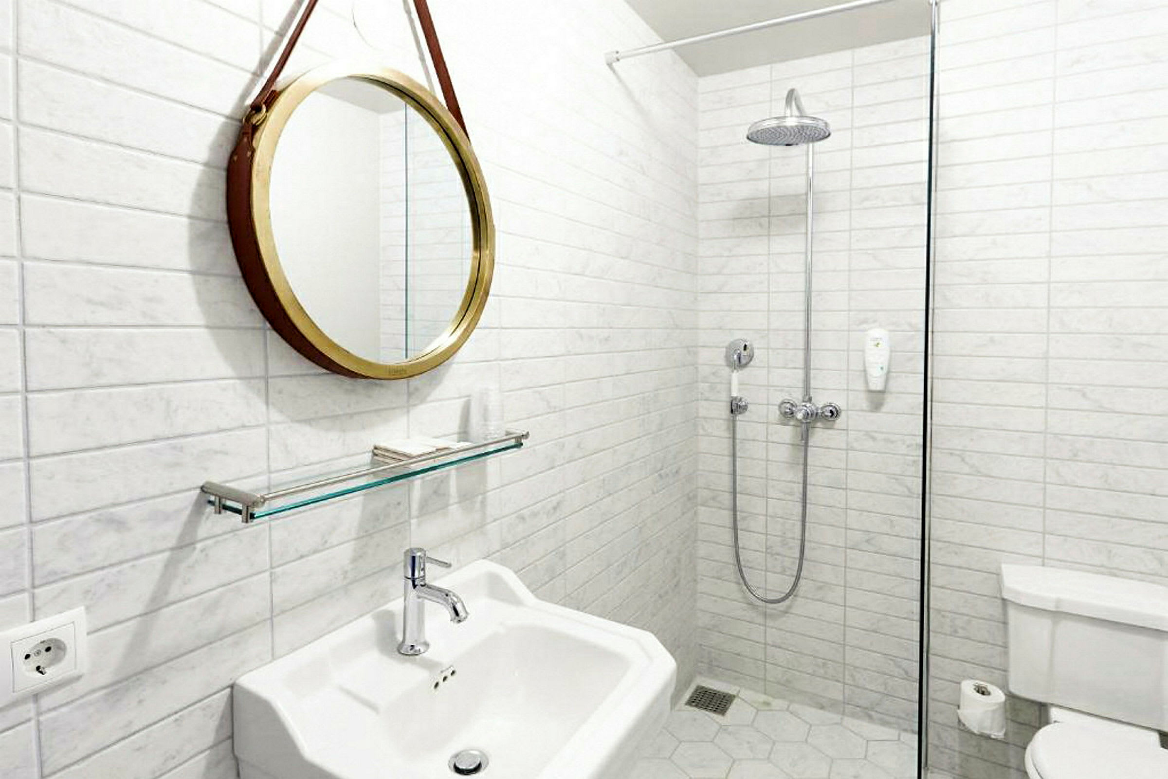 Et badeværelse med hvide fliser på væggene, et rundt spejl med guldramme, en håndvask, en bruser med glasvæg og et toilet. (AI-genereret tekst)