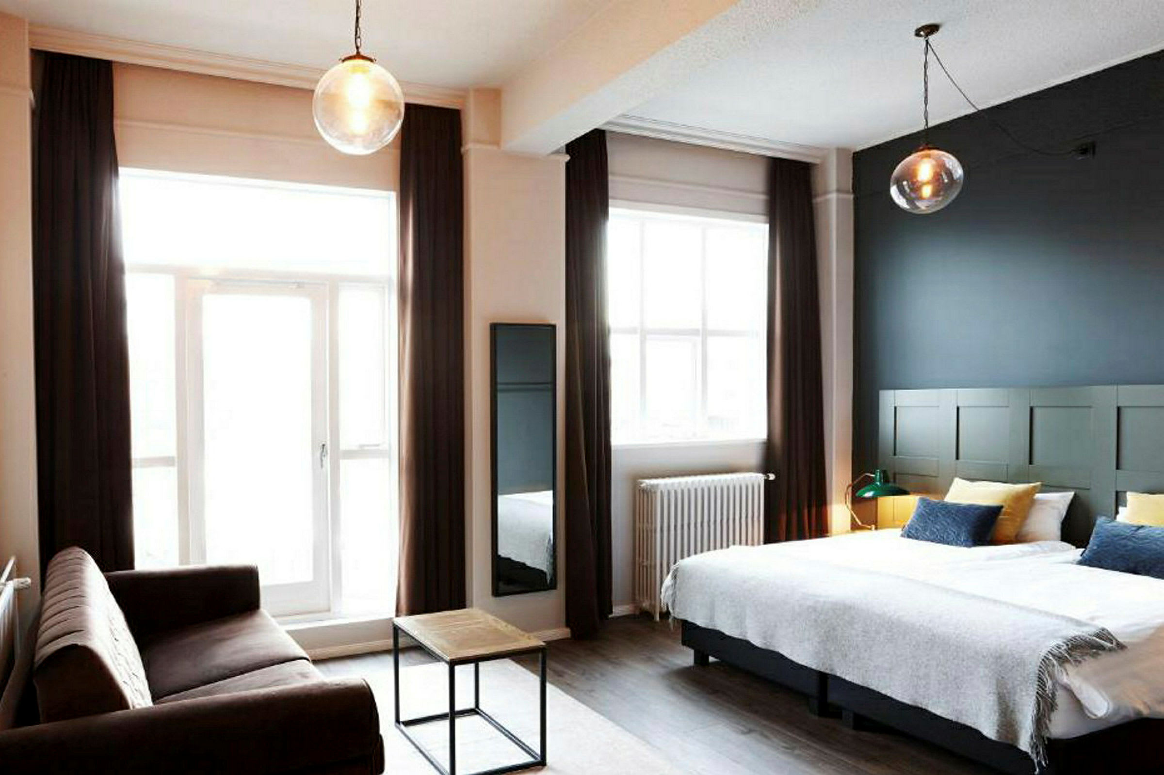 Et hotelværelse med en dobbeltseng, en sofa og et lille sofabord samt store vinduer med gardiner. (AI-genereret tekst)