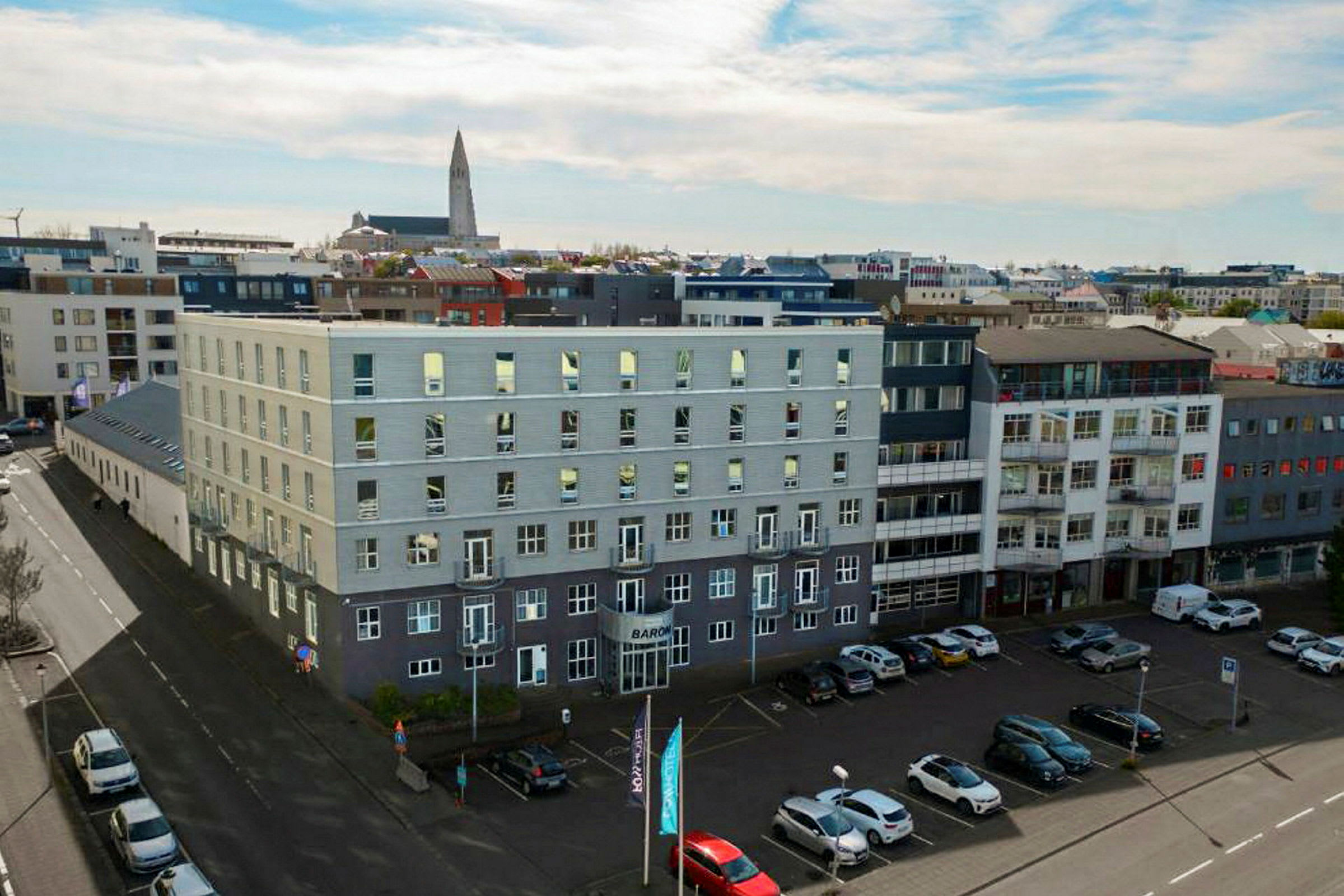 Flere moderne bygninger omgiver et åbent parkeringsområde, med biler parkeret på jorden og en kirke synlig i baggrunden. (AI-genereret tekst)