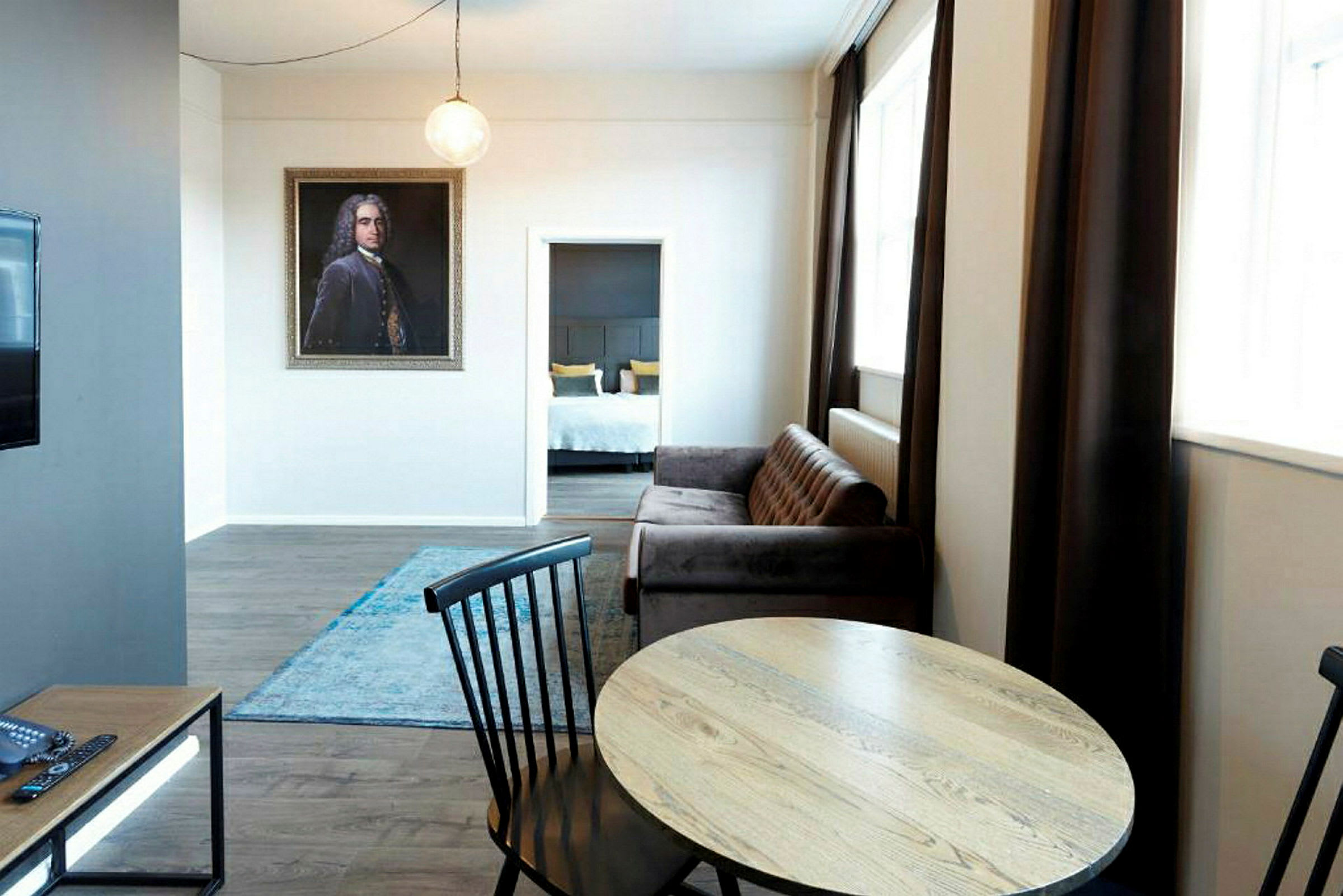 En stue med bord og stole foran en sofa, et tæppe på gulvet og en åbning til et soveværelse i baggrunden. (AI-genereret tekst)