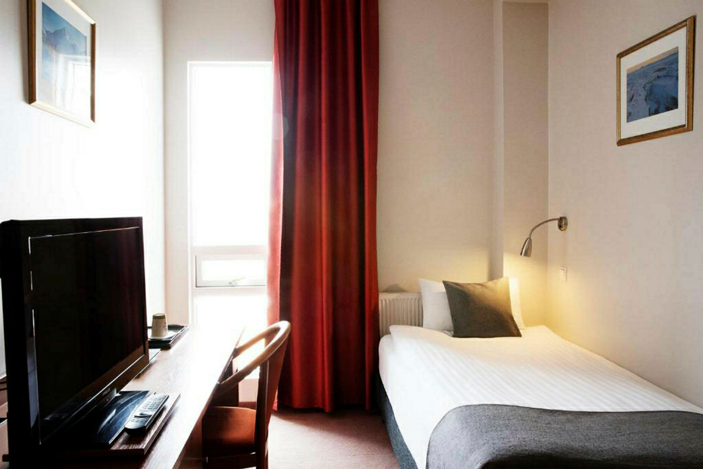 Et hotelværelse med en enkeltseng, skrivebord, TV og vindue med røde gardiner. (AI-genereret tekst)