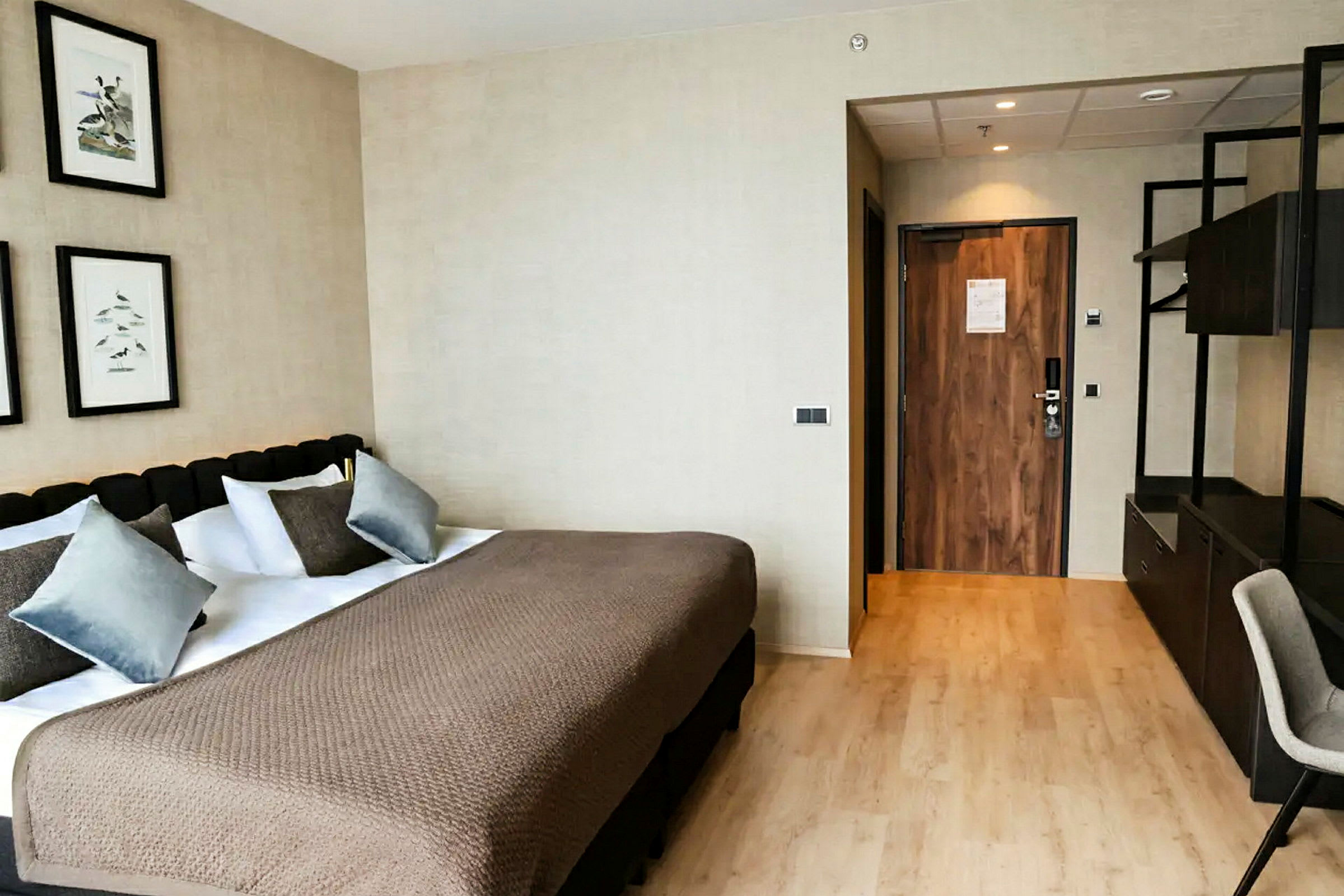 Et hotelværelse med en stor seng, flere puder og et beige sengetæppe. På væggen ovenfor hænger billeder, og en dør til højre fører ud. (AI-genereret tekst)