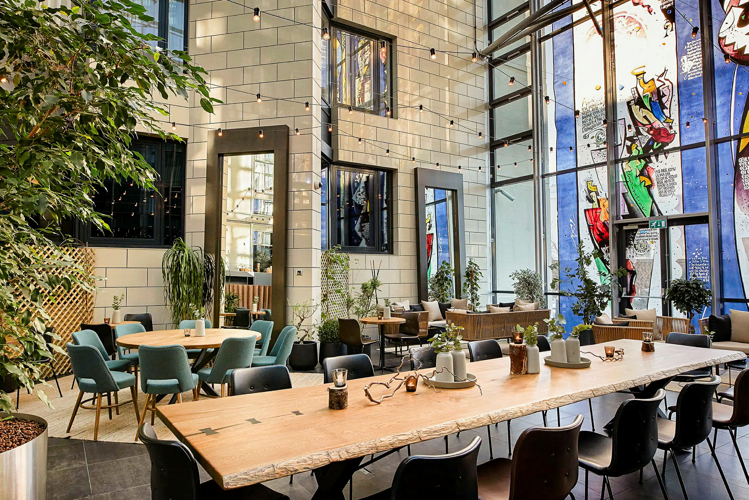 En indendørs lounge med store vinduer, siddepladser omkring flere borde og grønne planter placeret rundt omkring. (AI-genereret tekst)