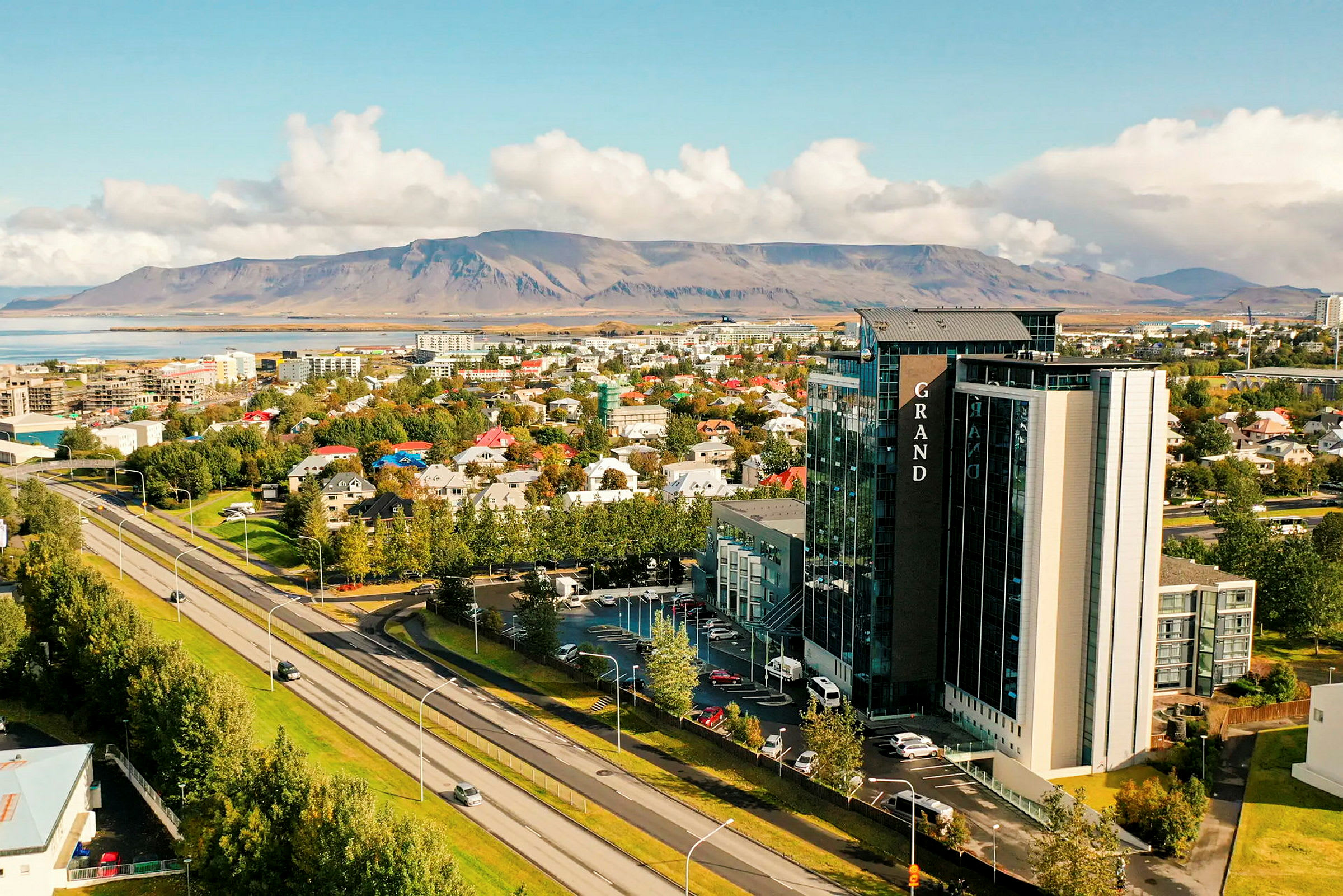 Et bymiljø med en motorvej i forgrunden, træer, bygninger og et større hotel. I baggrunden ses et bjerglandskab. (AI-genereret tekst)