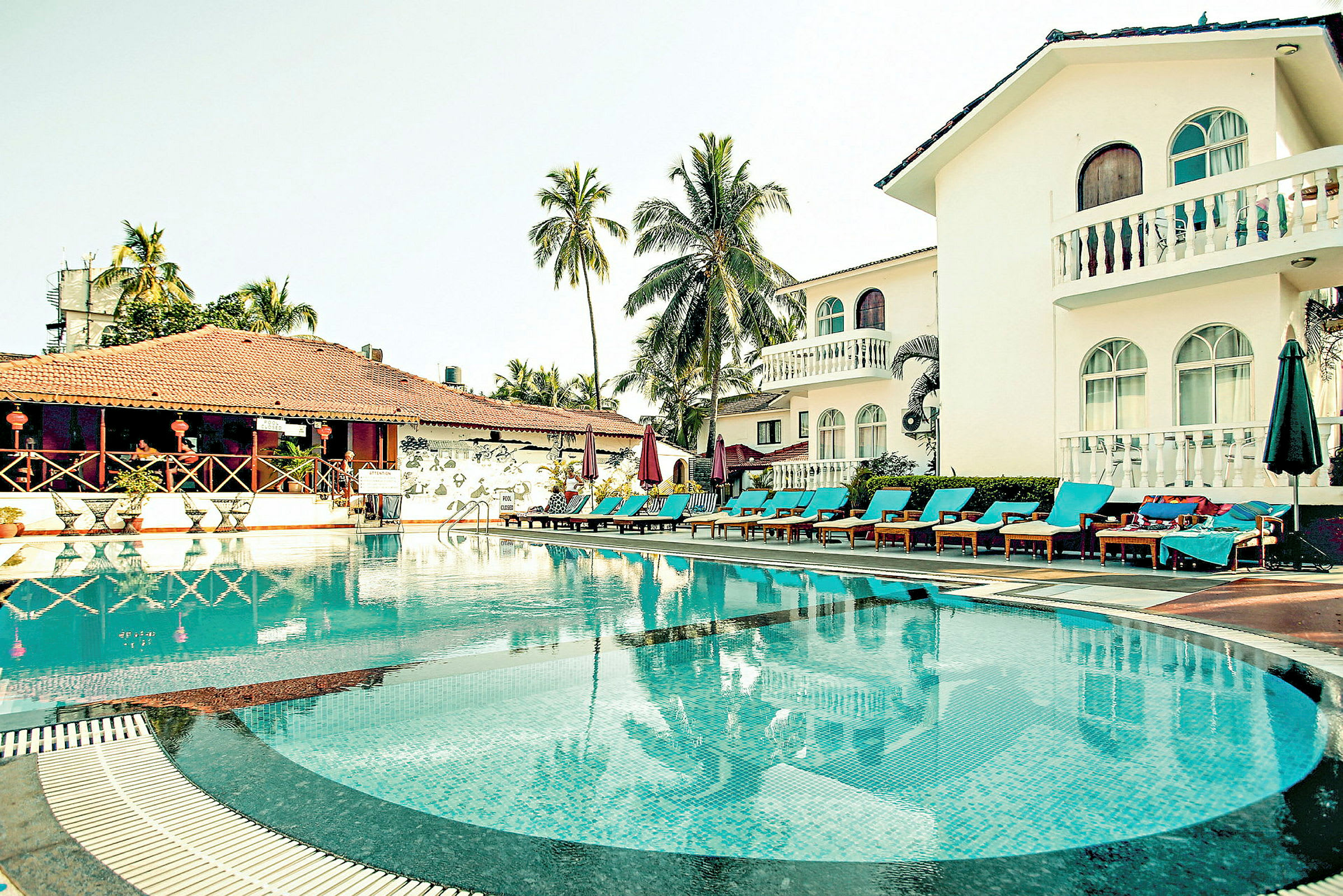 Billede af en pool omgivet af liggestole og bygninger med hvid facade. (AI-genereret tekst)