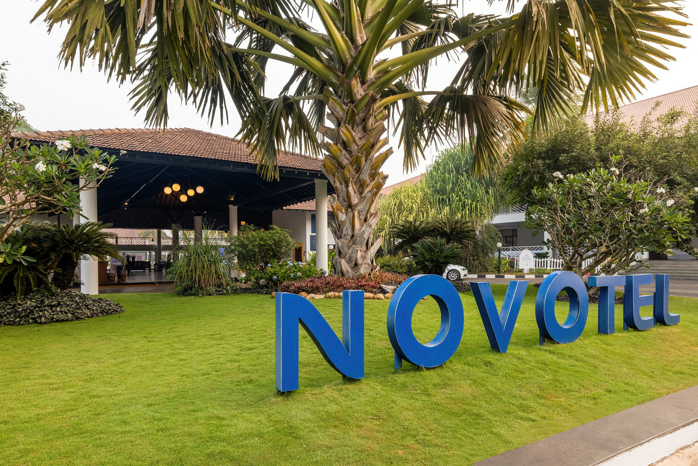 En have med en stor palme og grønt græs foran en bygning med et tag og et blåt skilt, der siger "Novotel". (AI-genereret tekst)