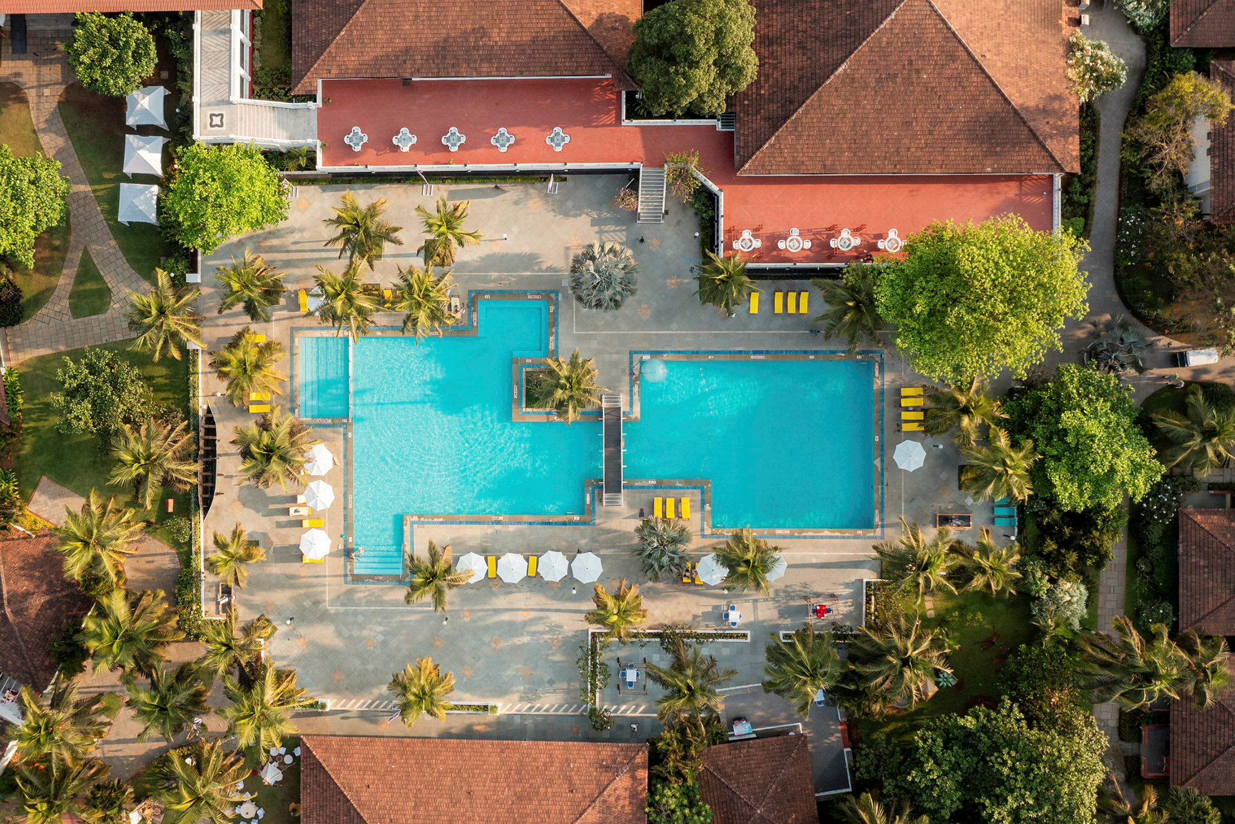 Luftfoto af et resort med to swimmingpools omgivet af palmer og bygninger med røde tage. (AI-genereret tekst)