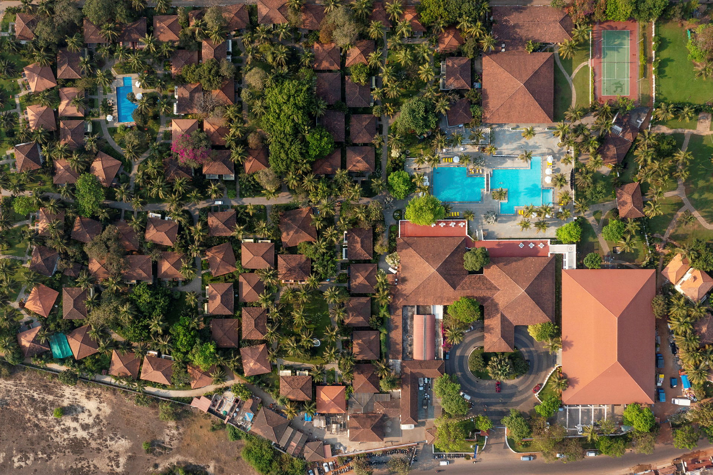 Et luftfoto, der viser et resort med terrakottafarvede tage, grønne områder, swimmingpools og tennisbaner. (AI-genereret tekst)