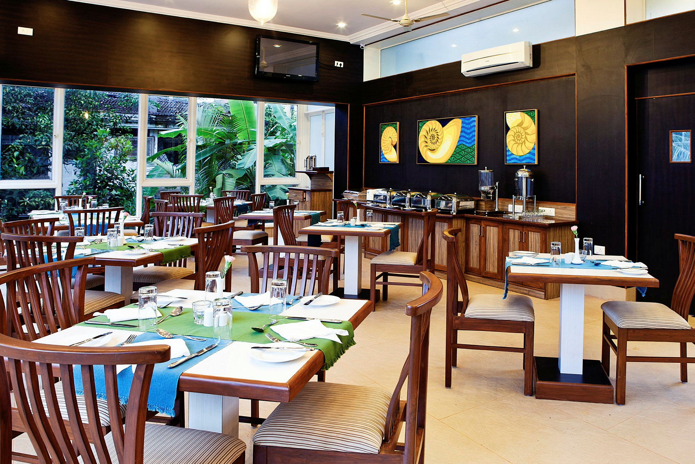 En restaurant med træborde og stole, farverige bordduge og dekorationer. Baggrunden viser en buffet med serveringscontainere. Vinduer giver udsigt til grønne planter. (AI-genereret tekst)