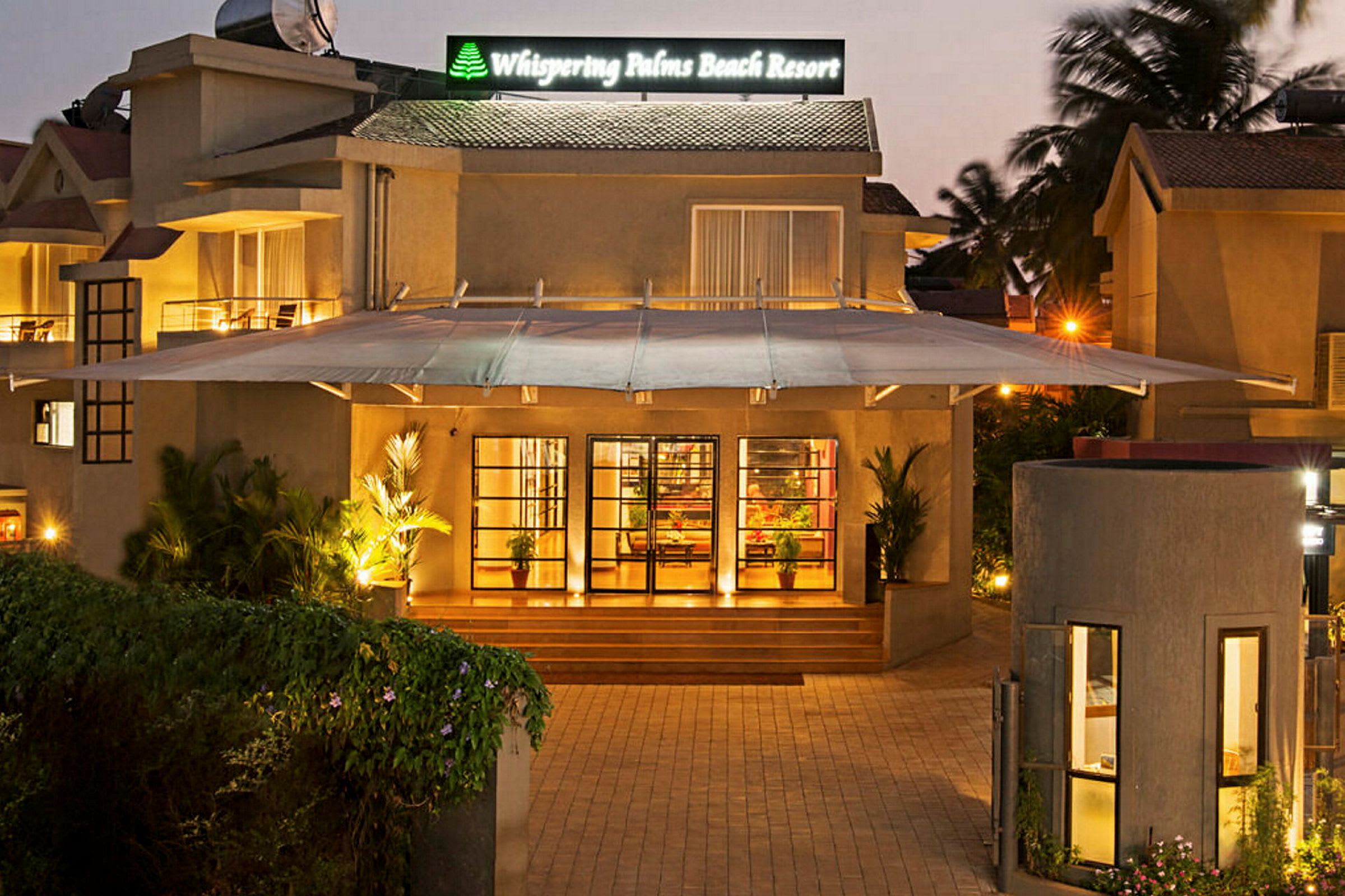 Indgangen til Whispering Palms Beach Resort med en belyst facade og grønne planter. (AI-genereret tekst)
