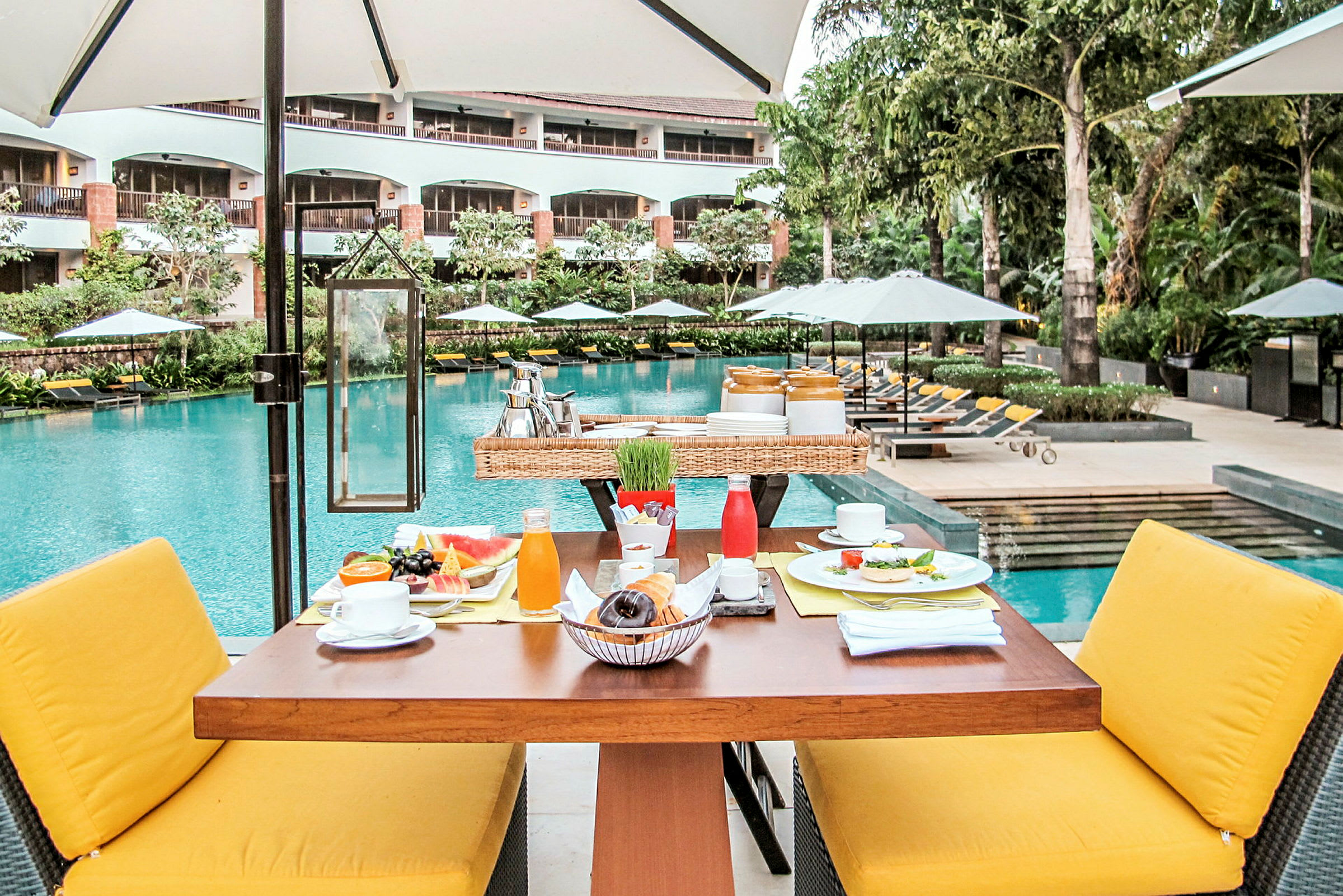 Morgenmad serveres på et bord med gule stole, omgivet af en swimmingpool og frodige haver. (AI-genereret tekst)