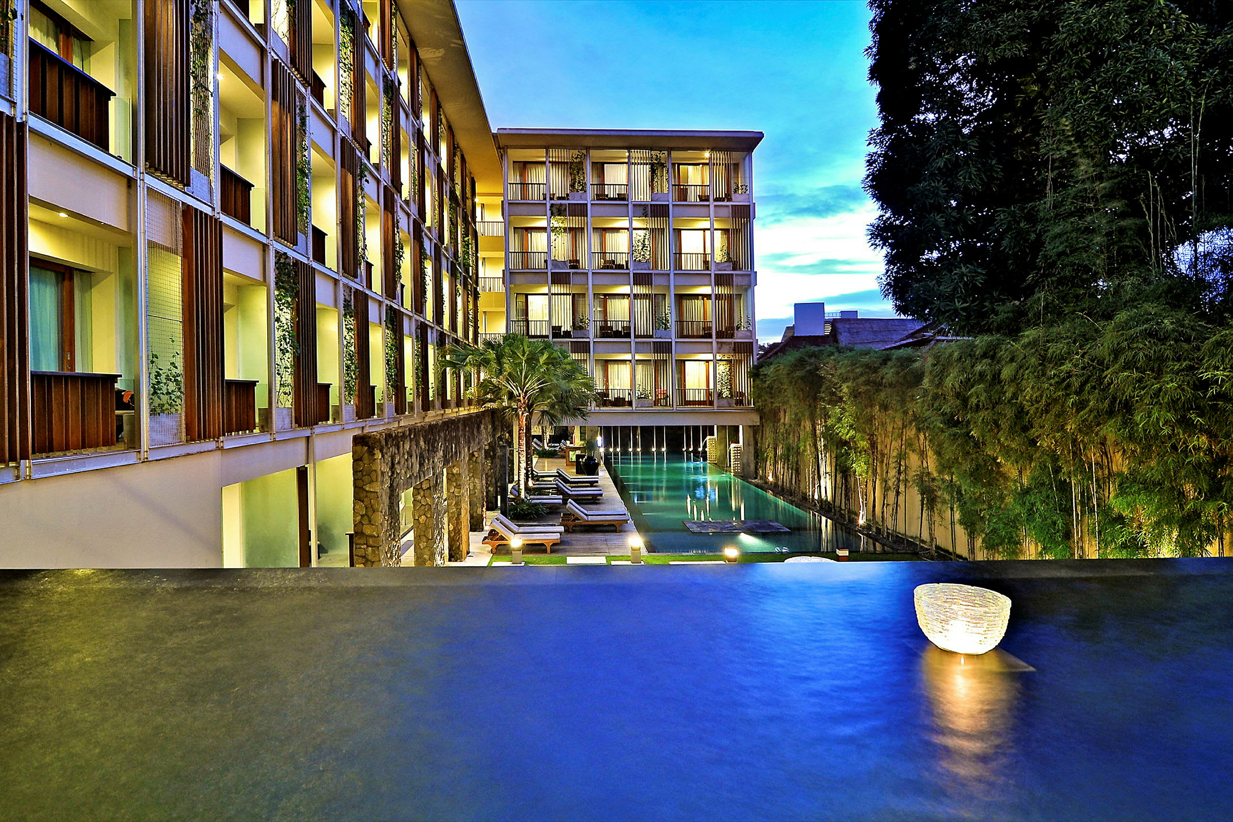 Billede af et moderne hotel med en stor pool omgivet af beplantning og flere etager med oplyste altaner. (AI-genereret tekst)