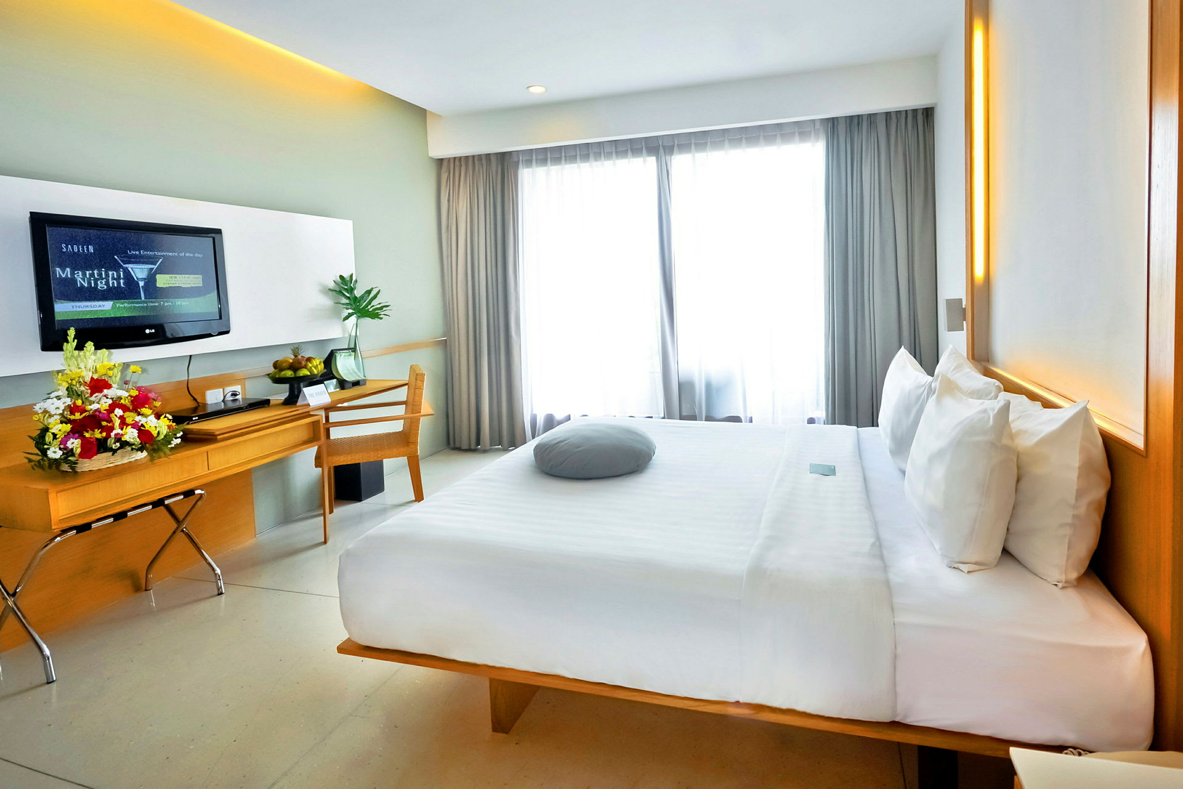 Et moderne hotelværelse med en stor komfortabel seng, skrivebord med blomsterarrangement og et TV på væggen. Vinduer med lyse gardiner lukker dagslys ind. (AI-genereret tekst)