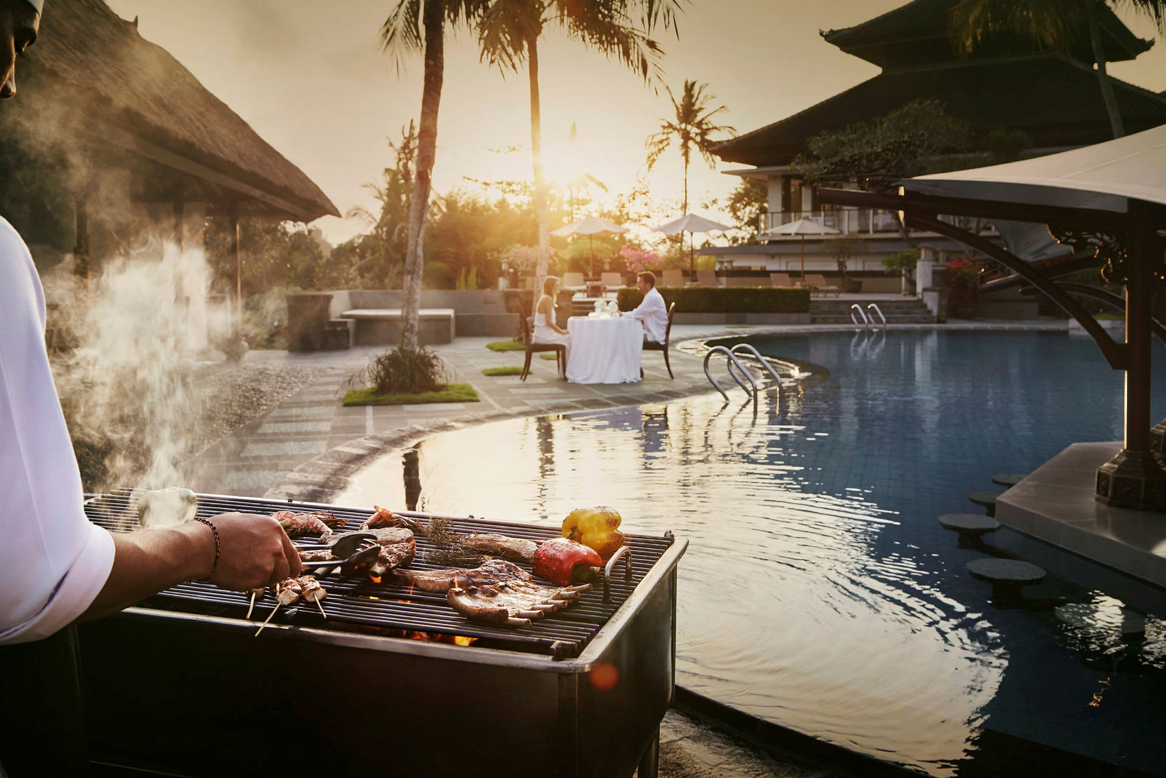En grill med madlavning tæt på en pool, hvor et par sidder ved et dækket bord. (AI-genereret tekst)
