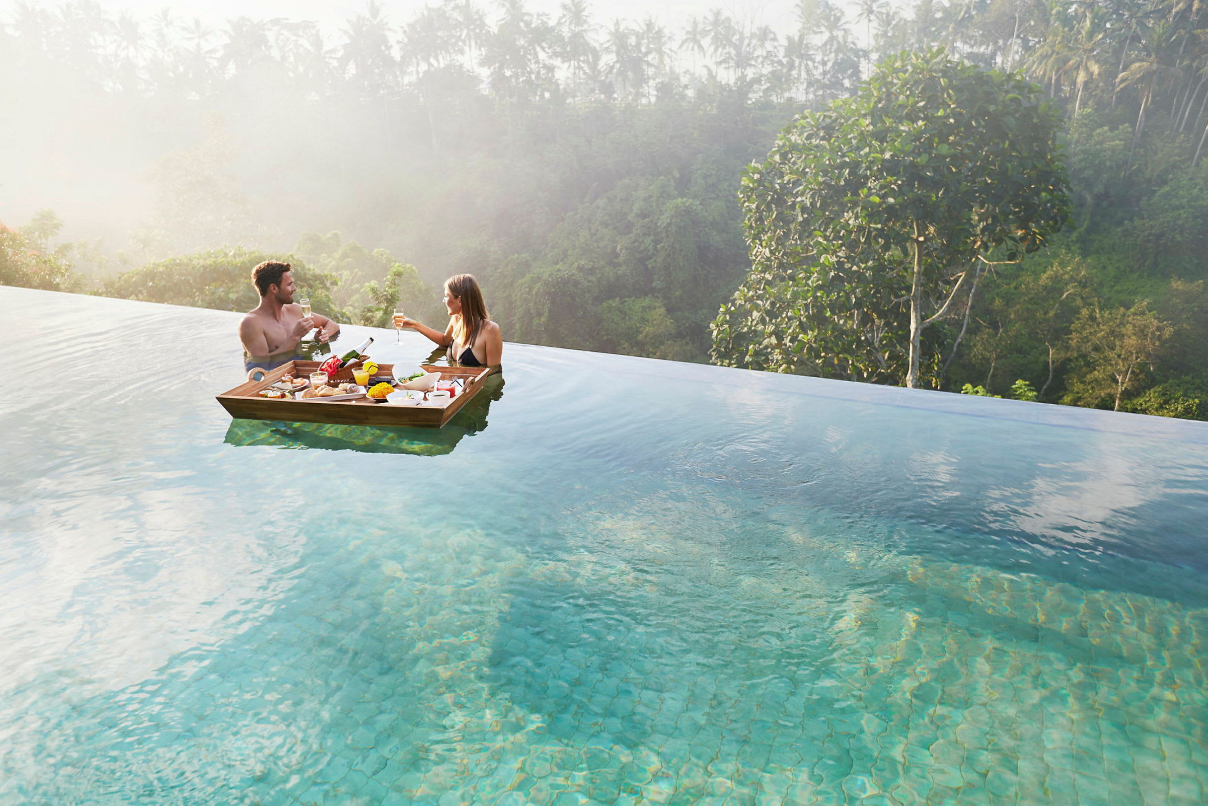 To personer nyder et måltid på en flydende bakke i en infinitypool, omgivet af frodig vegetation. (AI-genereret tekst)