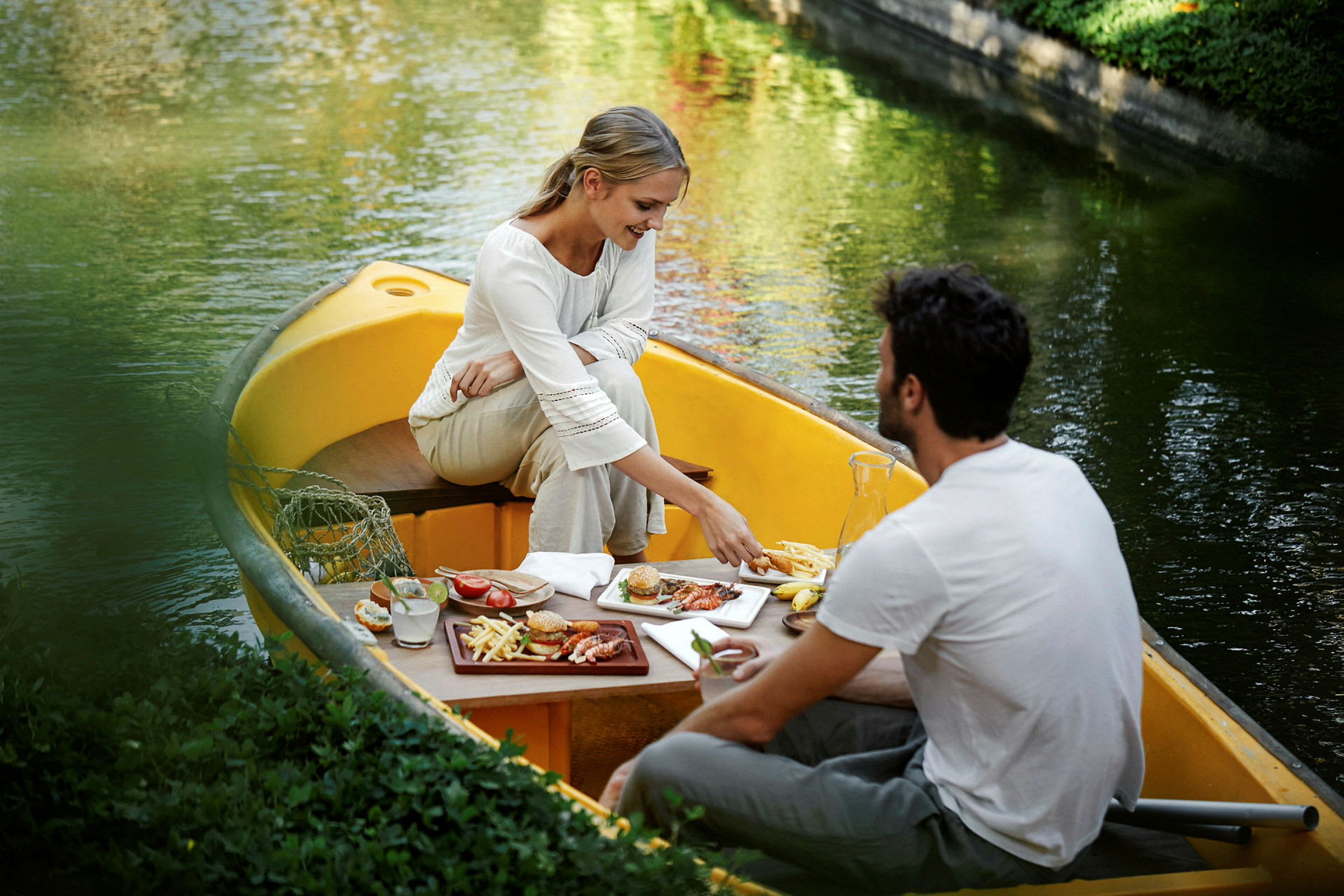 To personer sidder i en gul robåd på en rolig kanal og spiser et måltid ved et bord. (AI-genereret tekst)