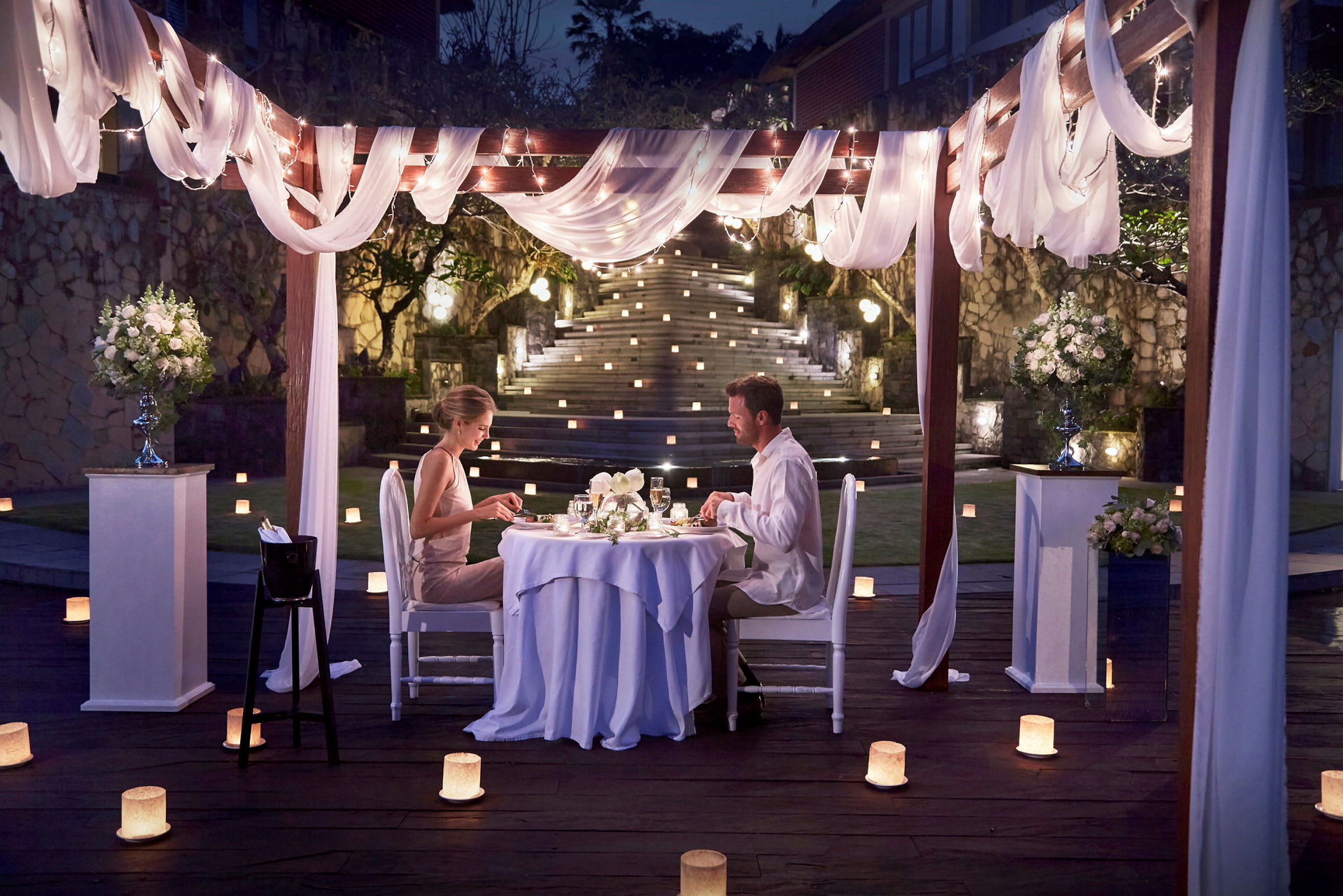 Et udendørs middagsbord under en elegant dekoreret pergola med lys og blomster. (AI-genereret tekst)