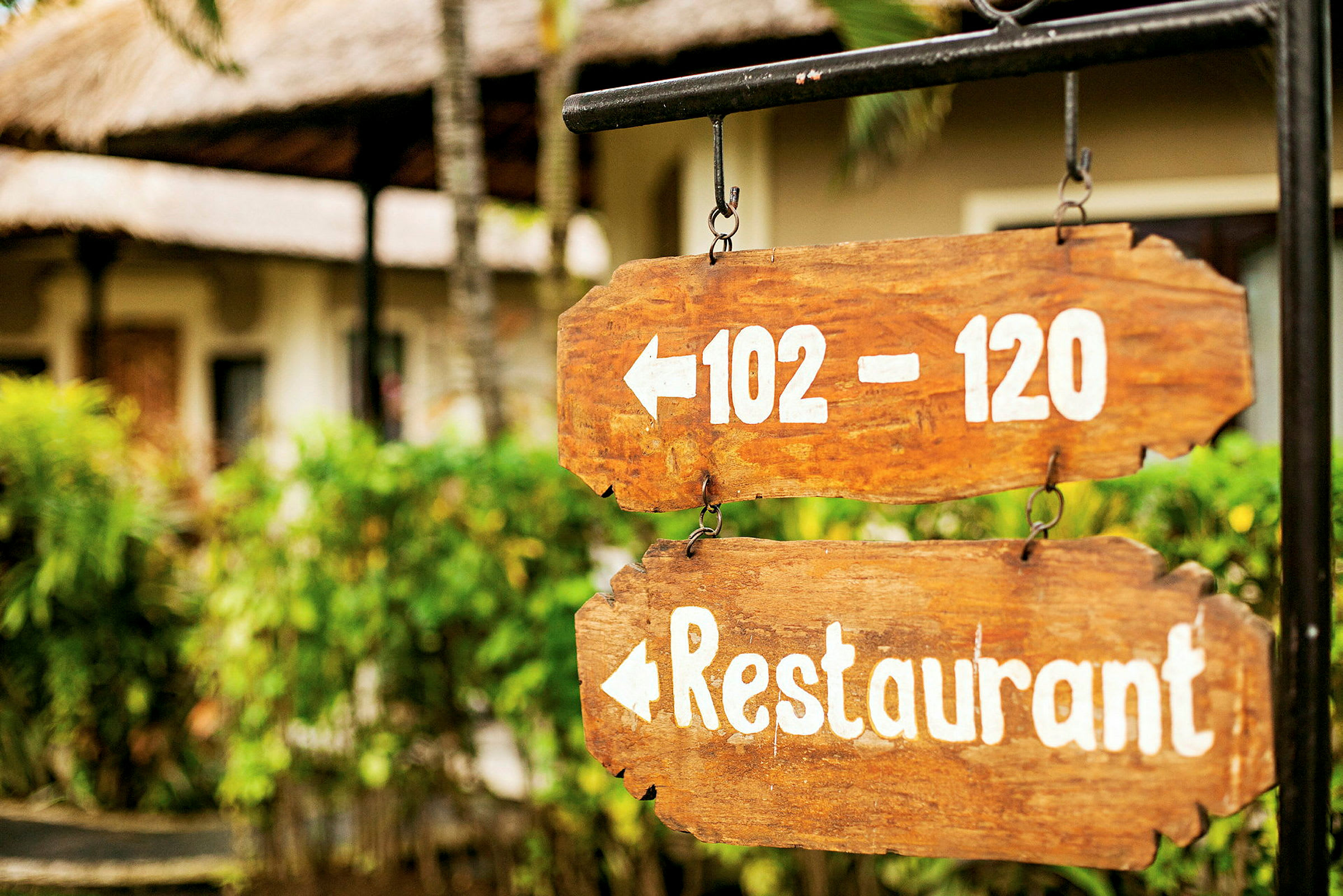 To træskilte med hvid tekst peger til venstre og viser tallene 102–120 og ordet restaurant. Baggrunden har grønne planter og bygninger. (AI-genereret tekst)