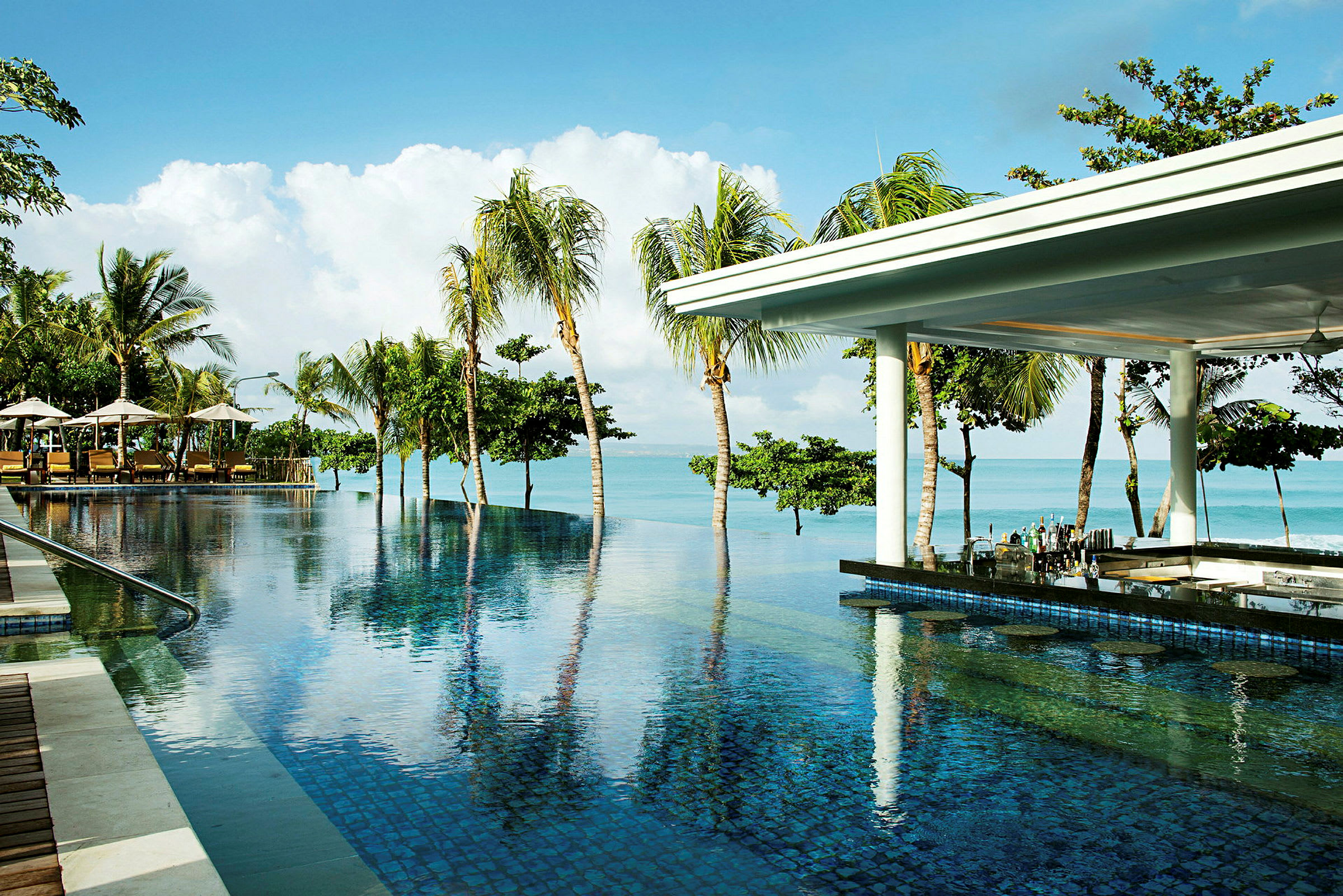 En infinitypool ved stranden med palmer i baggrunden og en bar under et tag ved poolen. (AI-genereret tekst)