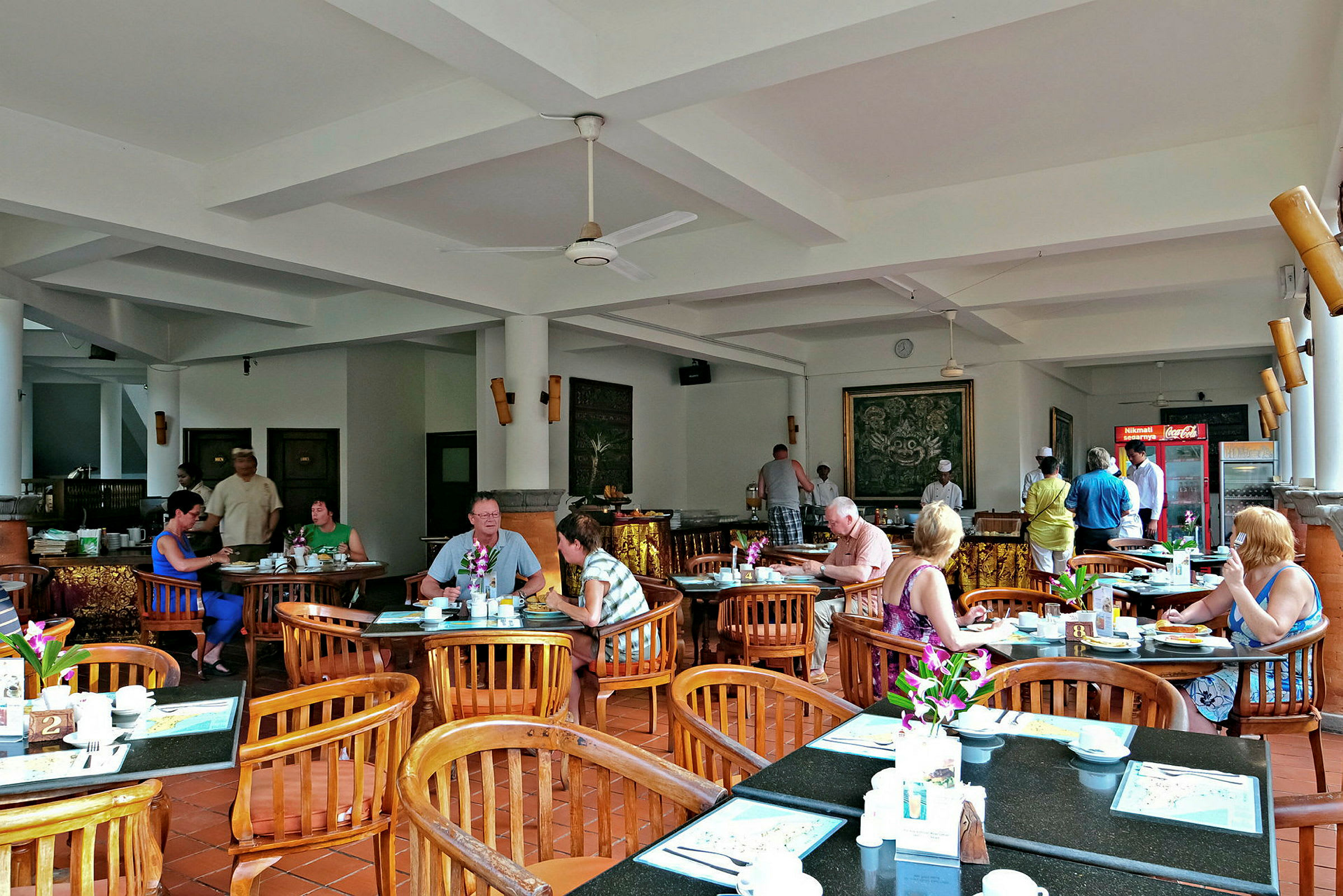 Et restaurantområde med borde og stole, hvor folk sidder og spiser. Indretningen er lys med hvide vægge og loft. (AI-genereret tekst)