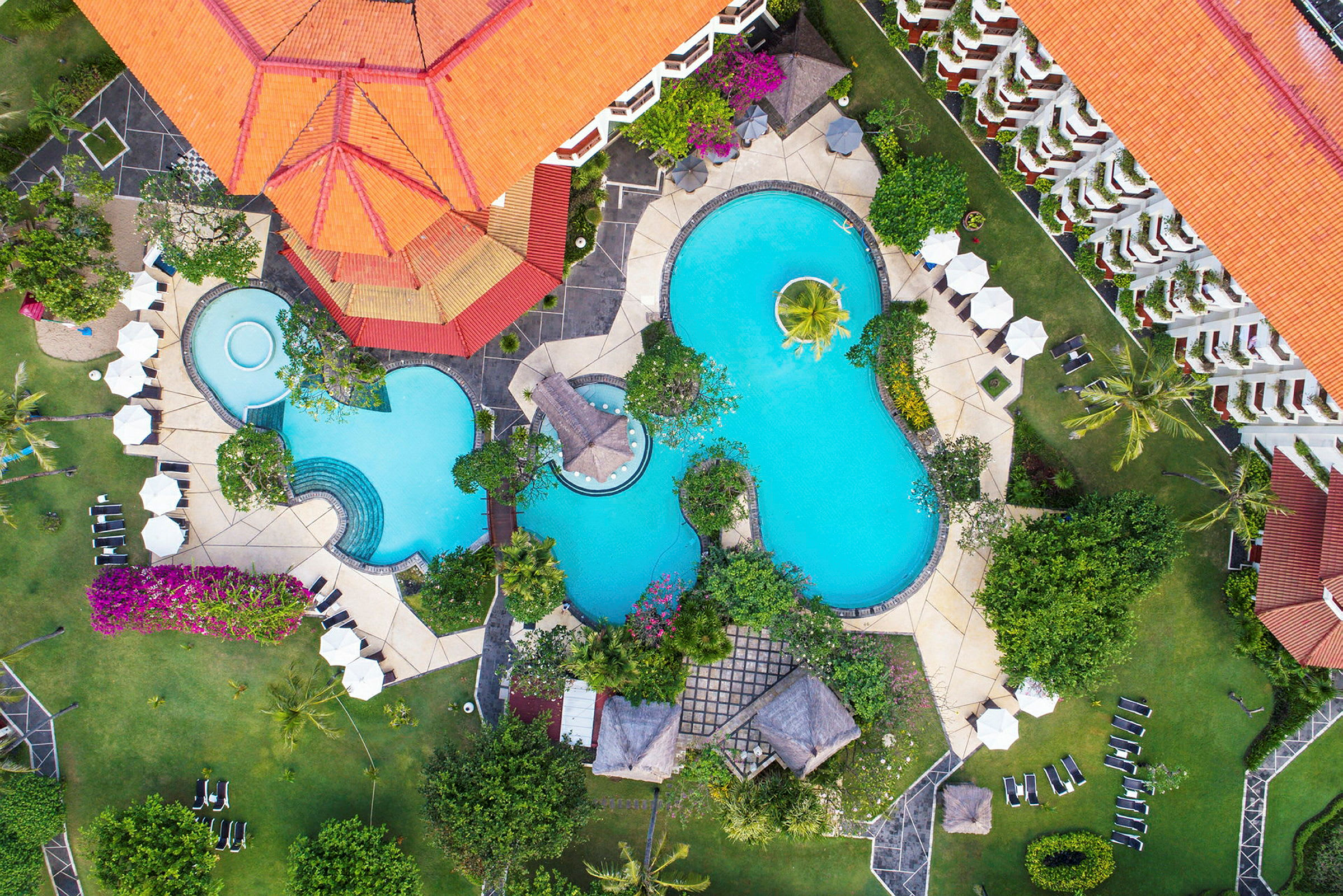 Luftfoto af en resort med flere swimmingpools omgivet af grønne haver og liggestole. (AI-genereret tekst)
