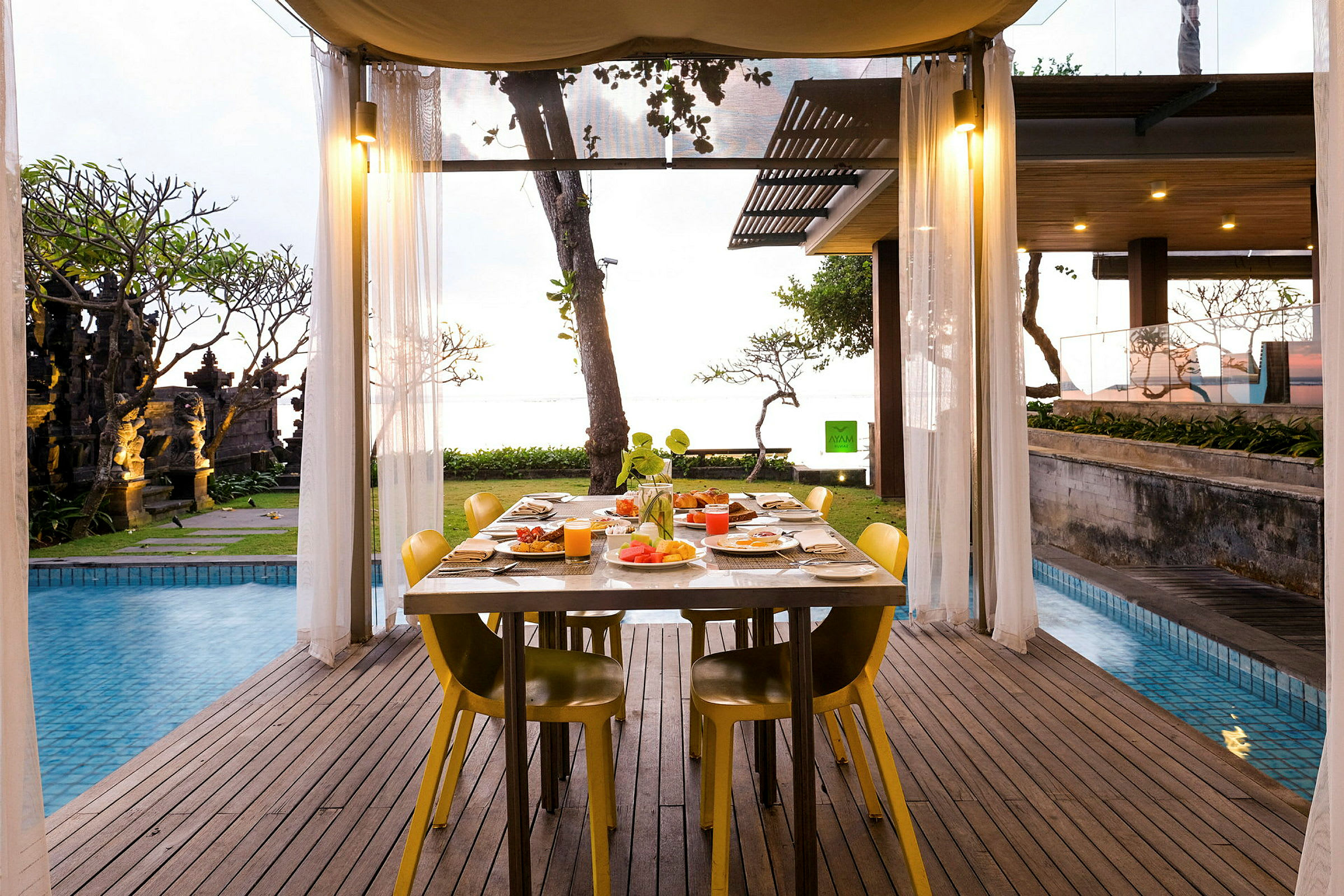 Et bord dækket til måltid står under en overdækket terrasse nær en pool med udsigt til haven og havet. (AI-genereret tekst)
