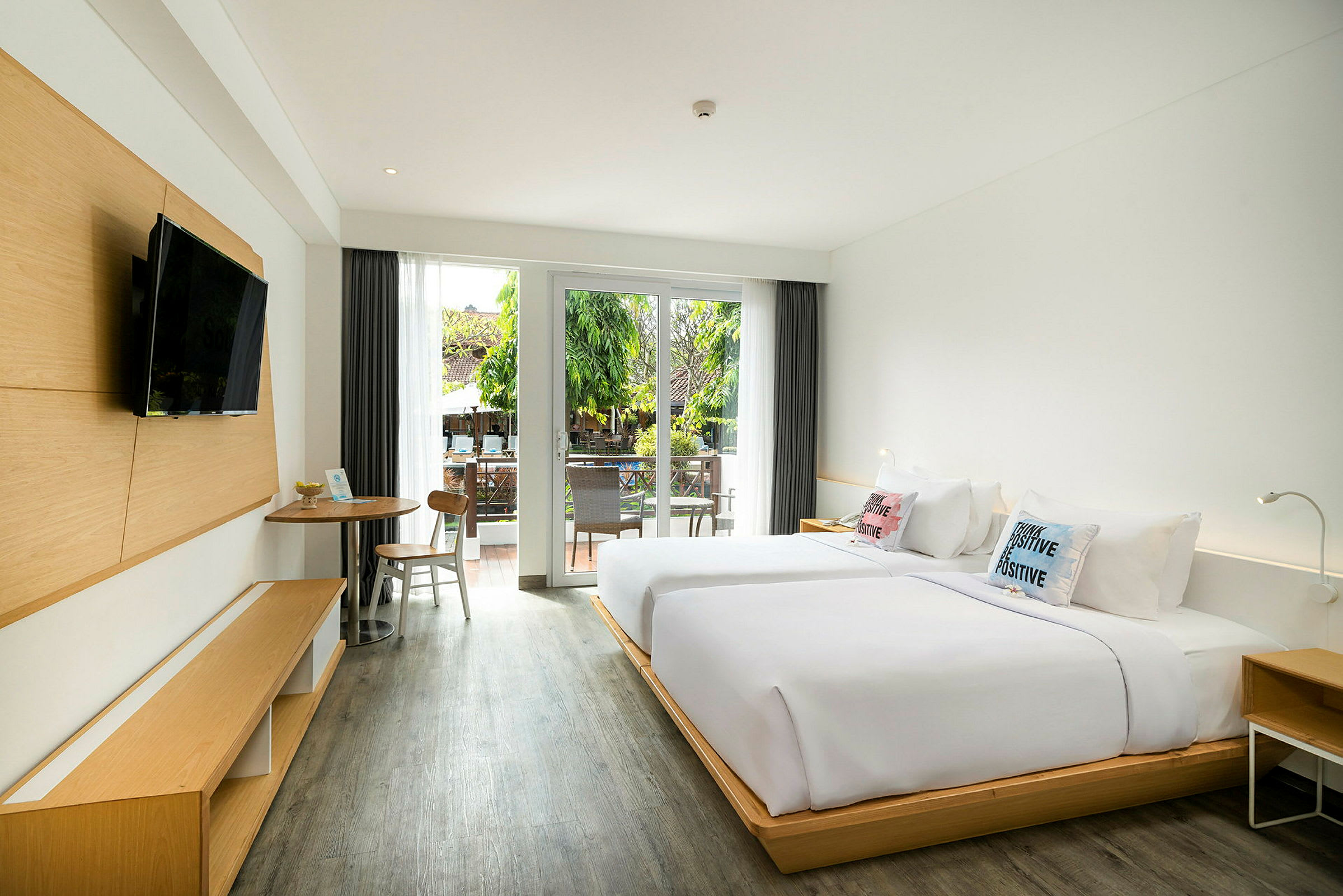 Et hotelværelse med to senge, et bord med stole og et moderne TV på væggen. Værelset har store glasdøre, der fører ud til en balkon med udsigt over grønt. (AI-genereret tekst)