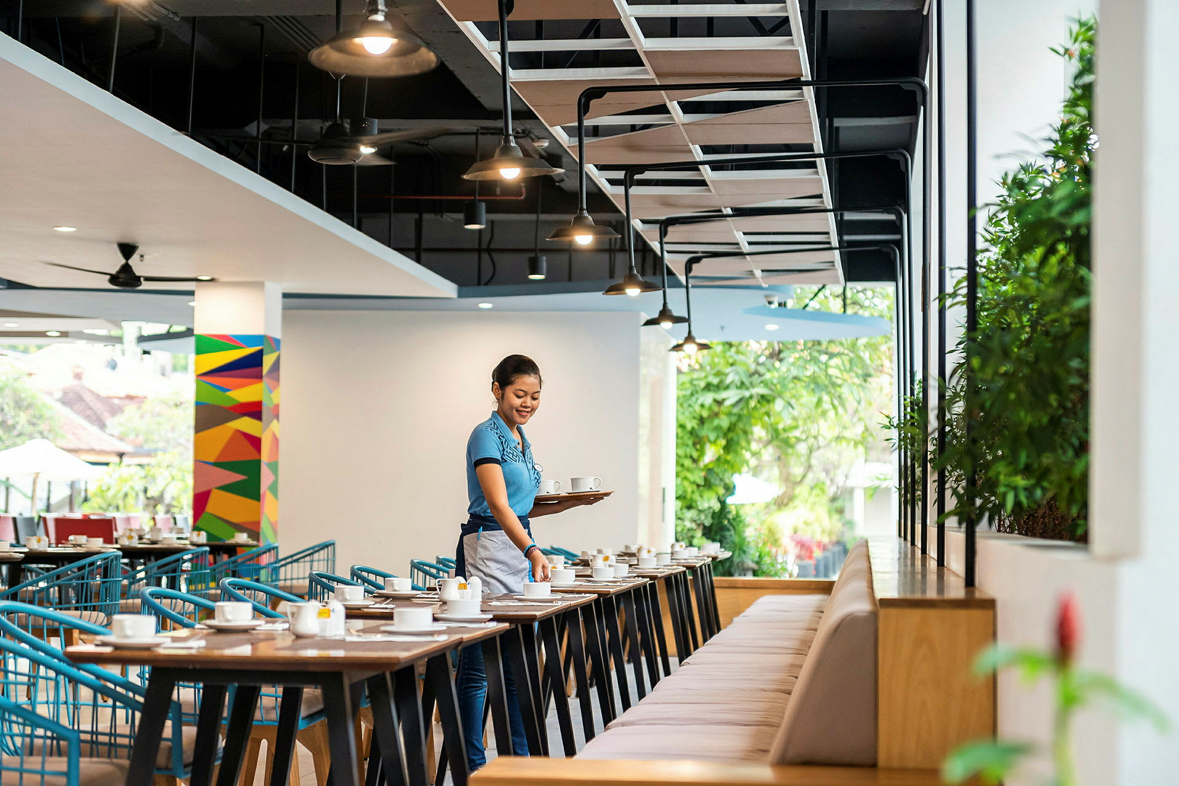 En restaurantlokale med borde og stole, hvor en person bærer en bakke med kopper. (AI-genereret tekst)