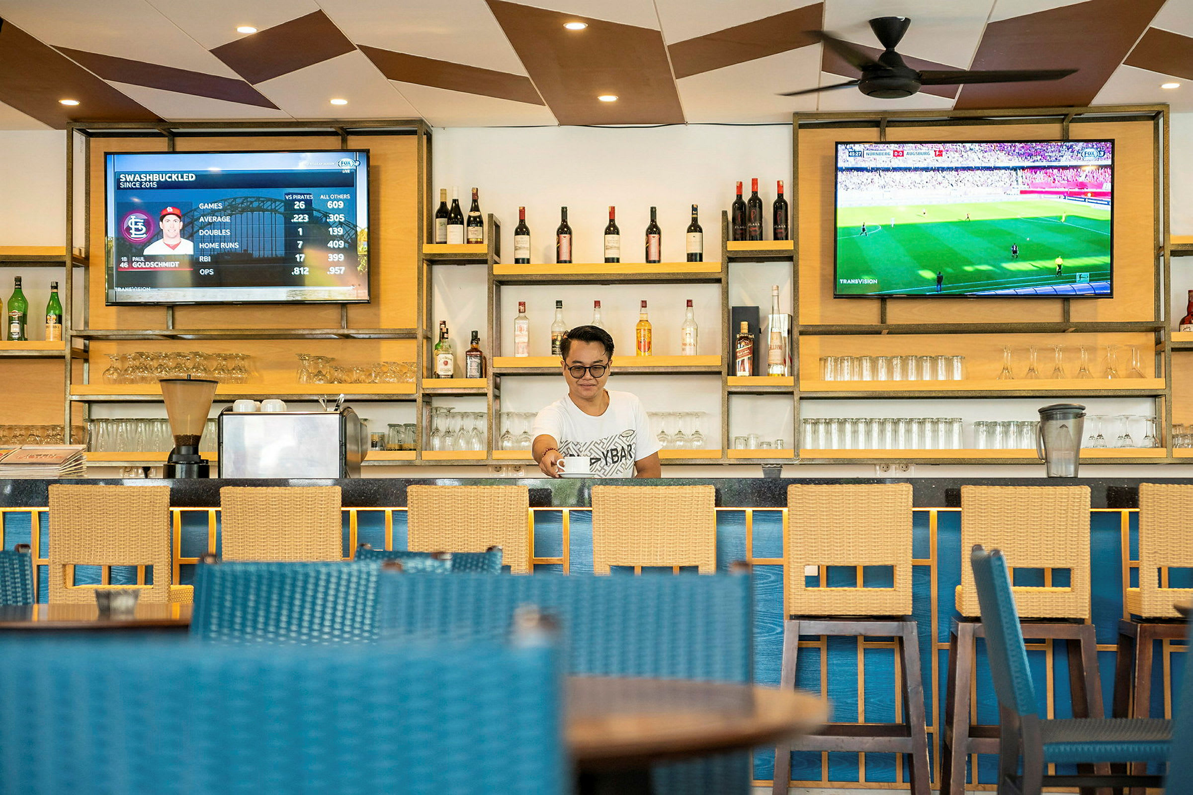En bar med elegante træmøbler, flasker og glas på hylder samt to skærme der viser sportsdata og en livekamp. (AI-genereret tekst)