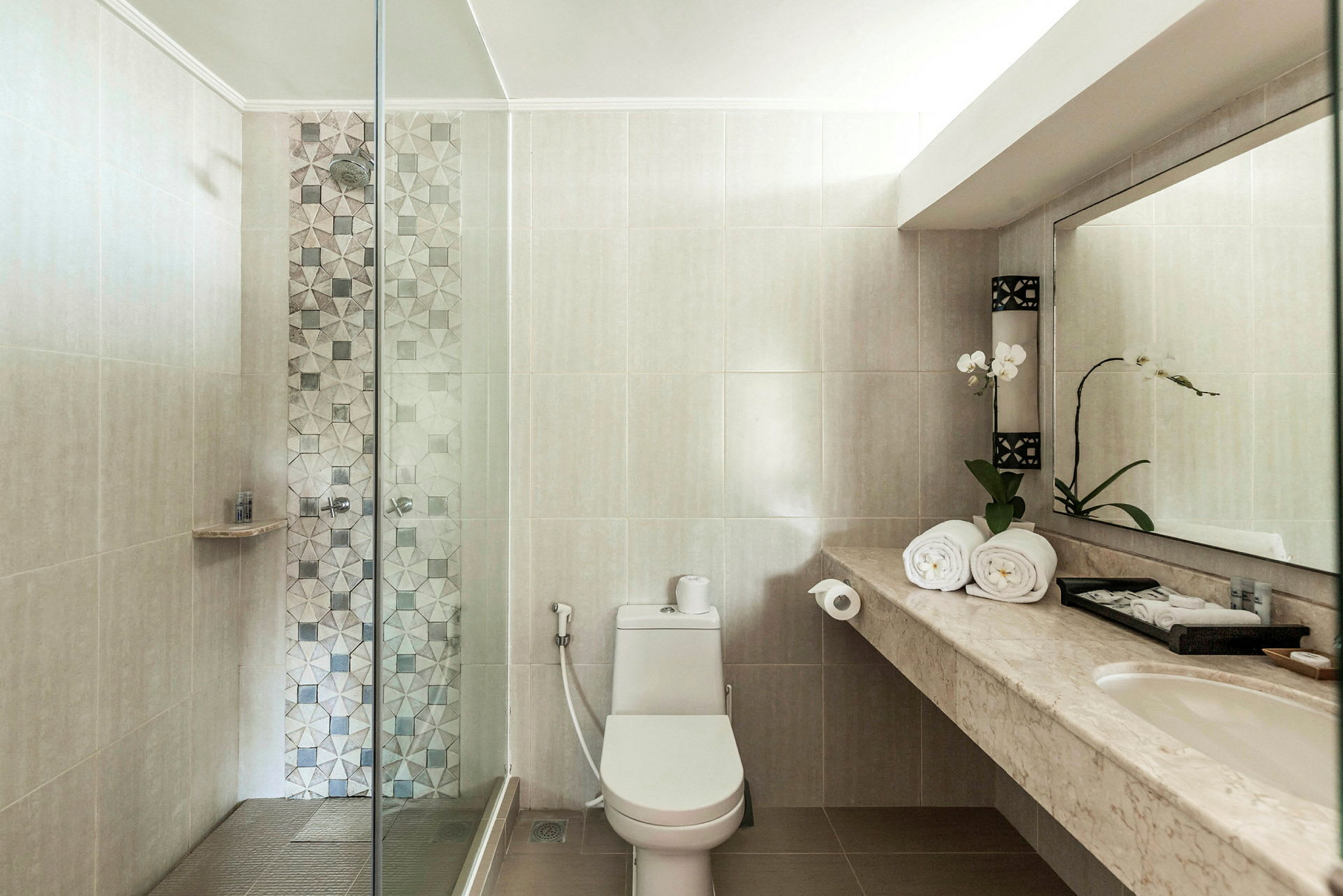 Et badeværelse med brusekabine, toilet og en marmordisk med håndvask, håndklæder og en blomst. (AI-genereret tekst)