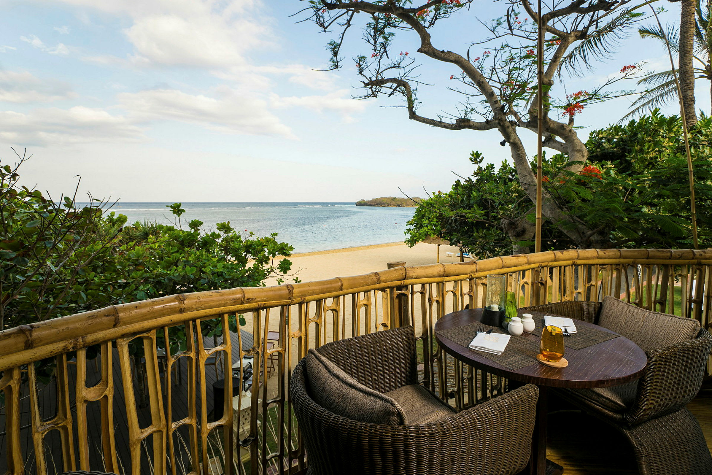 Et bord med kurvestole står på en balkon med udsigt over en sandstrand og havet, omgivet af frodige træer. (AI-genereret tekst)