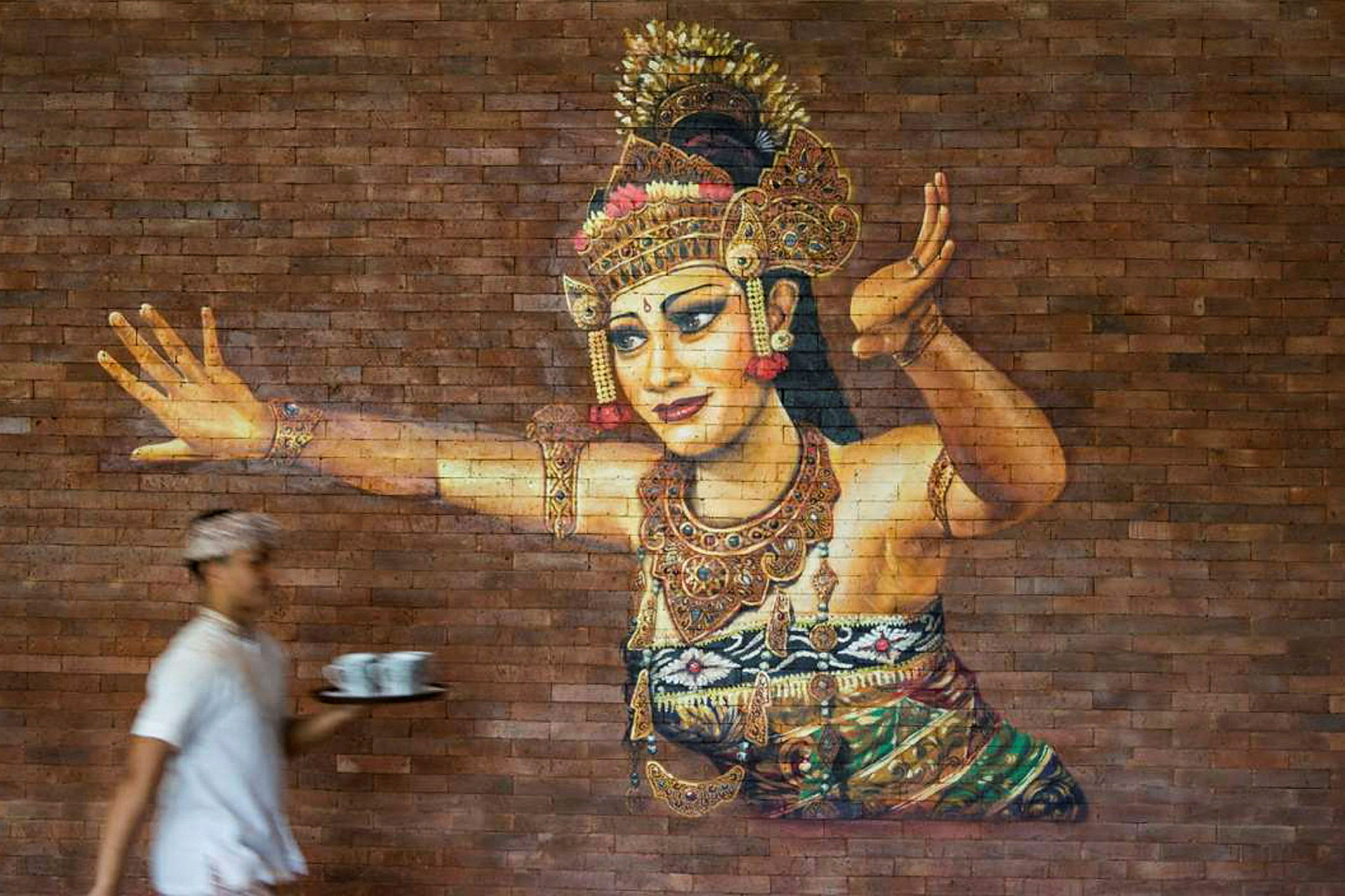 Et kunstværk på en murstensvæg, der viser en traditionel danser i udsmykkede klæder og smykker. En person i forgrunden holder en bakke med kopper. (AI-genereret tekst)