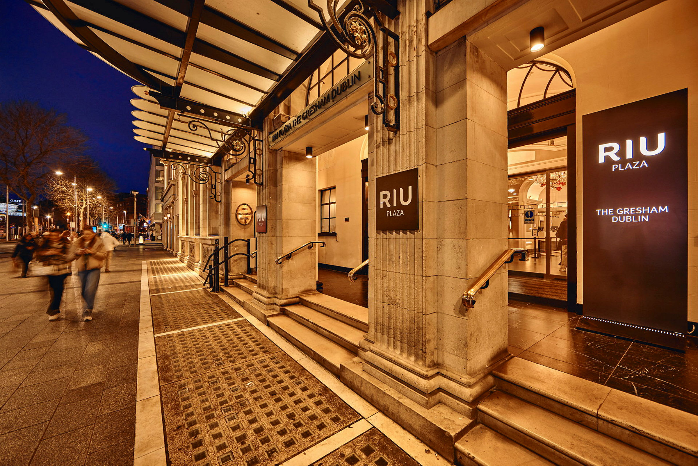 Hotel RIU Plaza Gresham Dublin indgang med oplyste skilte, aftenstemning. (AI-genereret tekst)