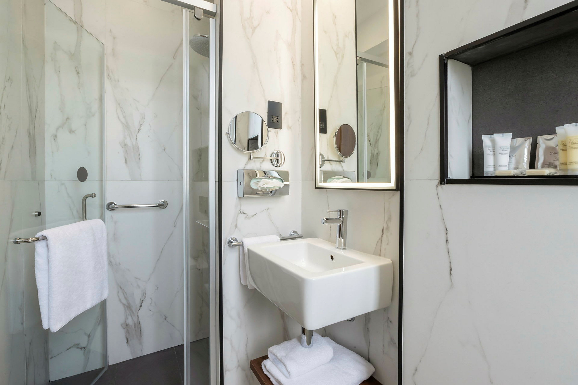 Foto af et badeværelse med brusebad, håndvask, spejl og toiletartikler på en hylde. (AI-genereret tekst)