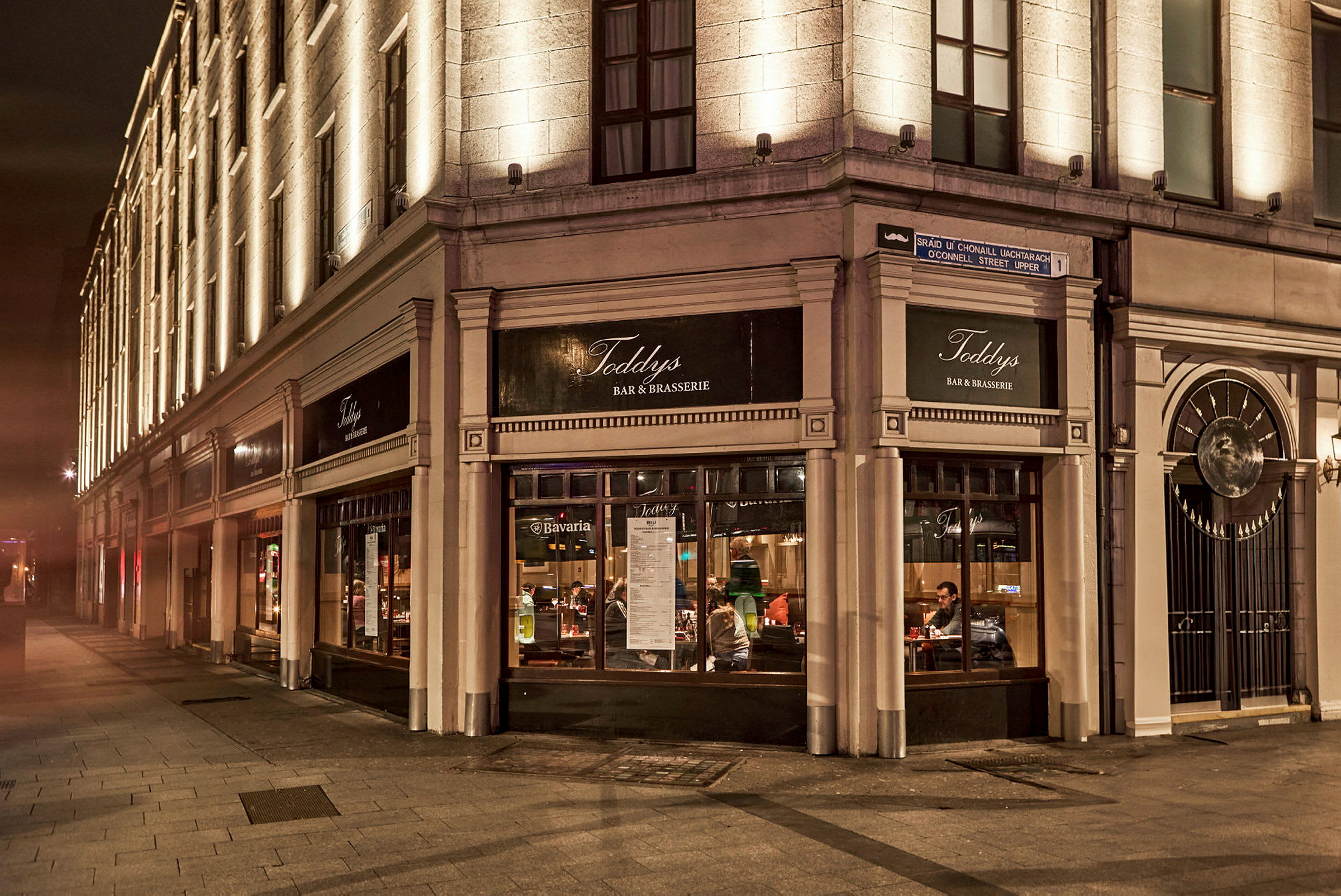 En bygning af sten med belyste facade og en restaurant markeret med skilte, der viser Toddy's Bar & Brasserie. (AI-genereret tekst)