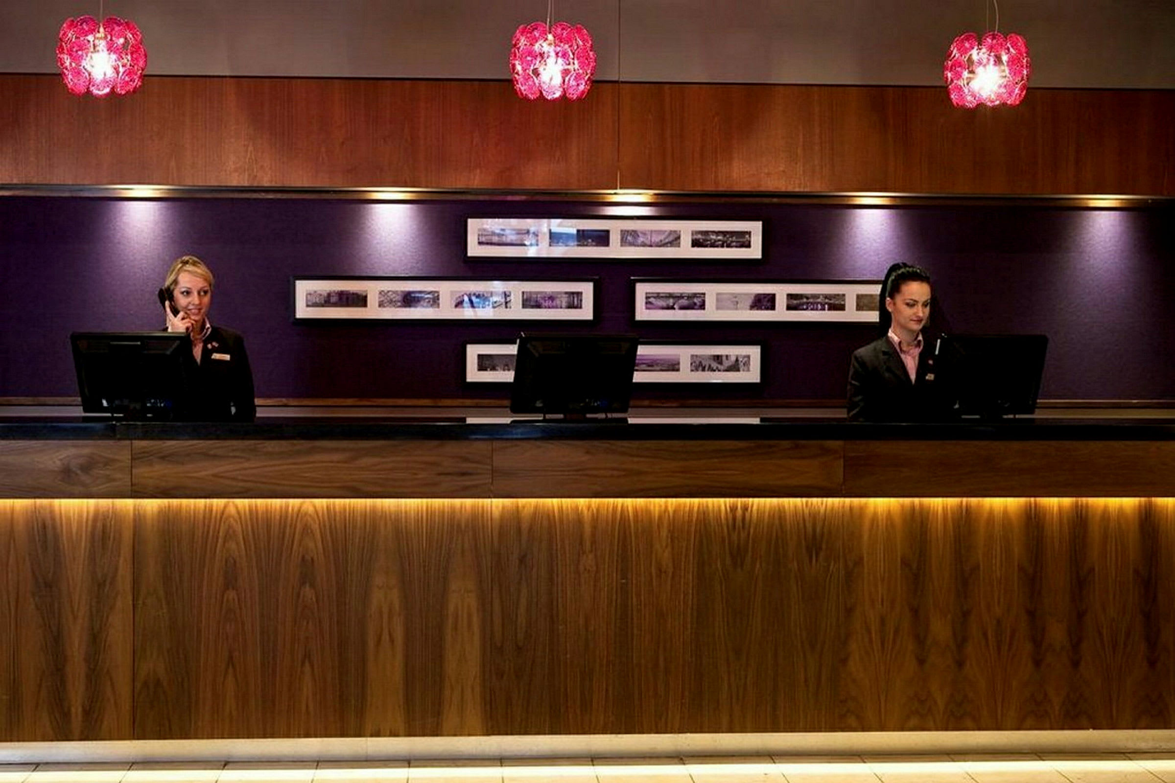 To receptionister står bag en receptionsdisk med computerskærme og telefoner. Baggrunden har en polstret væg med indrammede billeder. (AI-genereret tekst)
